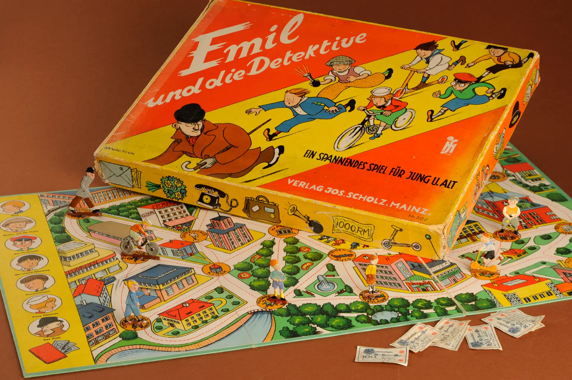 Brettspiel "Emil und die Detektive" mit Spielfiguren auf einem bunten Spielplan mit Häusern und rotem Spielkarton mit Titel und Illustrationen.