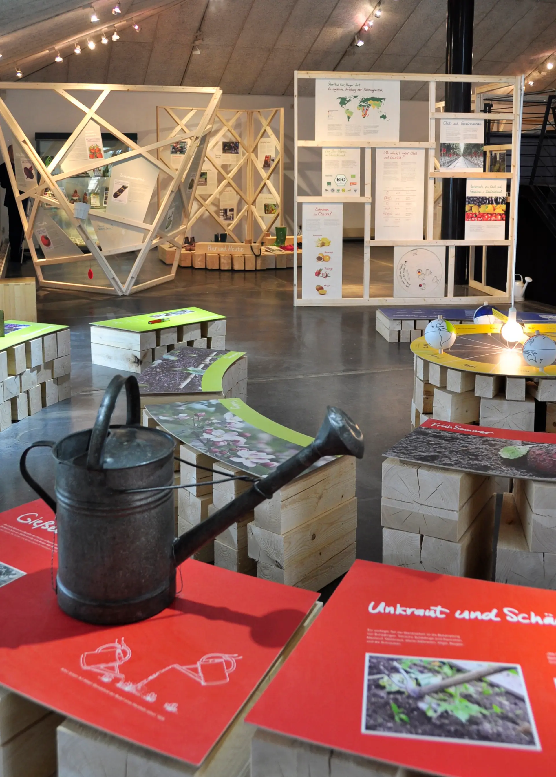 Ausstellung mit hölzernen Sitzmöbeln, Informationswänden und einer Gießkanne auf einem roten Tisch mit Infotafeln.
