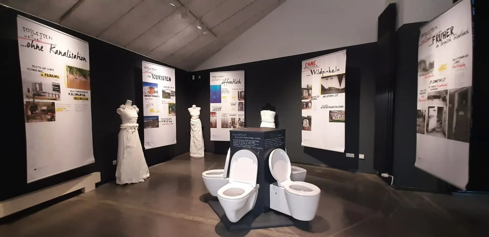 Blick in eine Ausstellung mit Texttafeln an den Wänden, drei Toiletten mit geöffneten Deckeln in der Mitte und drei weißen Papierkleidern auf Ständern.