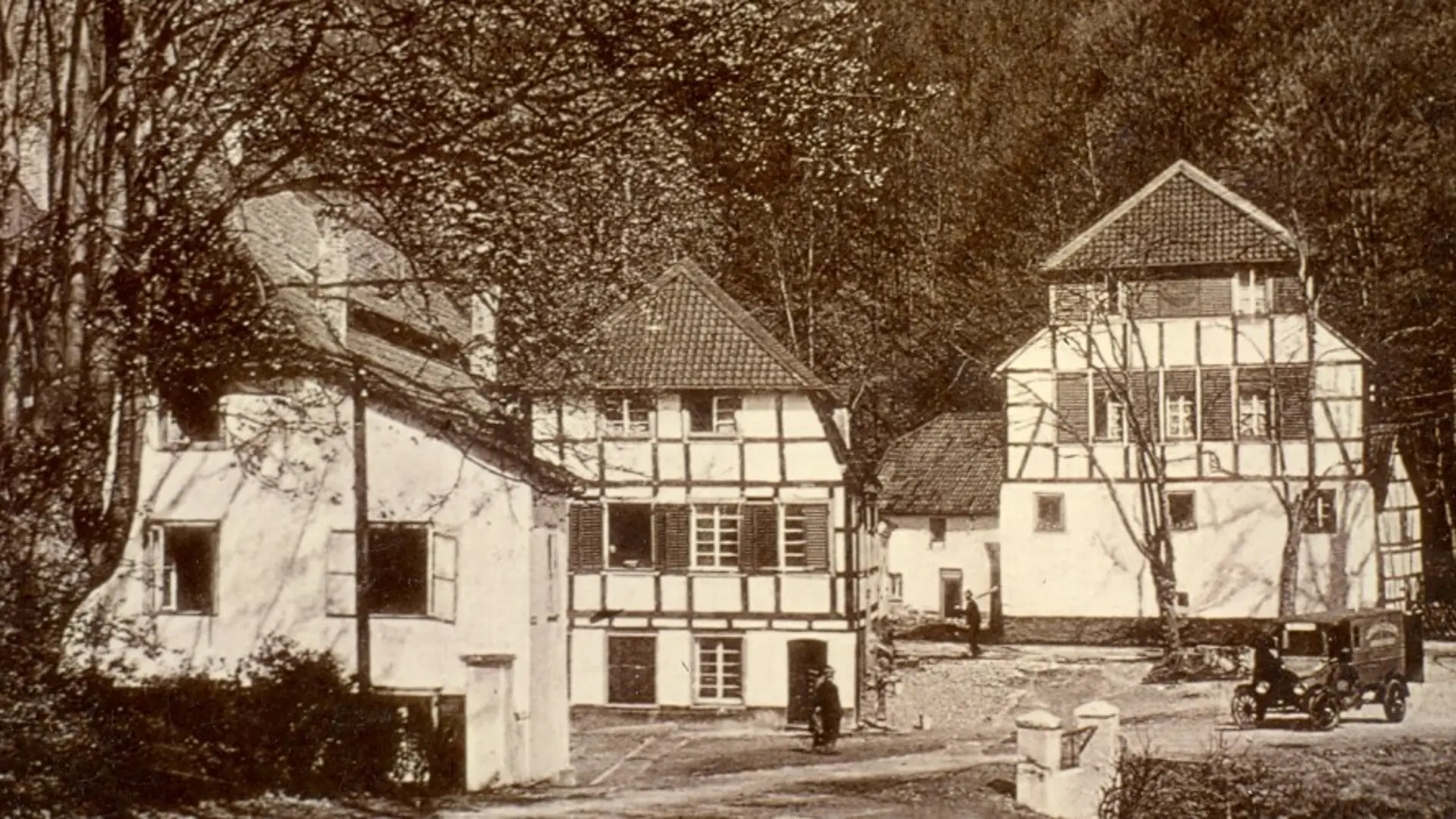 Historische Aufnahme in Sepia von mehreren Fachwerkgebäuden, die von Bäumen umsäumt werden.