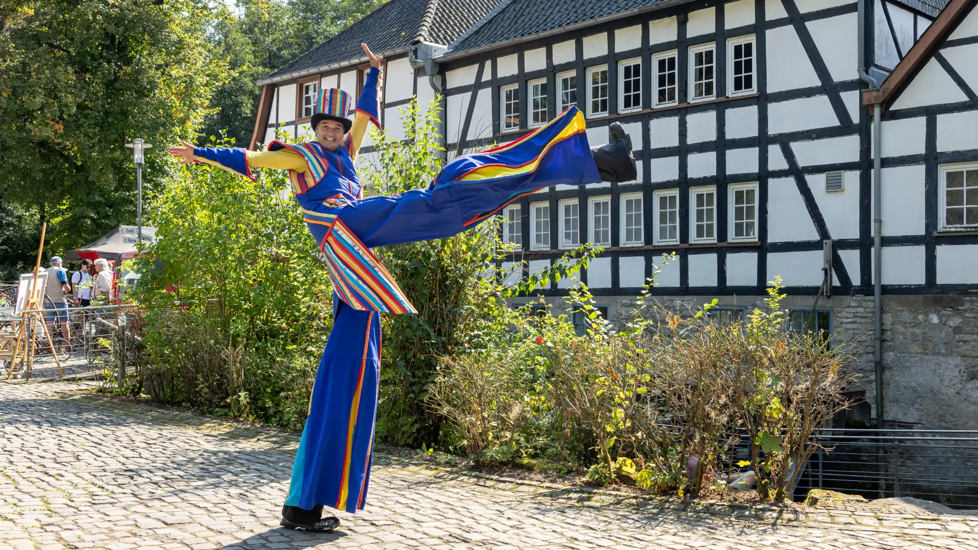 Ein Mann mit Stelzen in bunter Kleidung hebt ein Bein, im Hintergrund ein Fachwerkgebäude