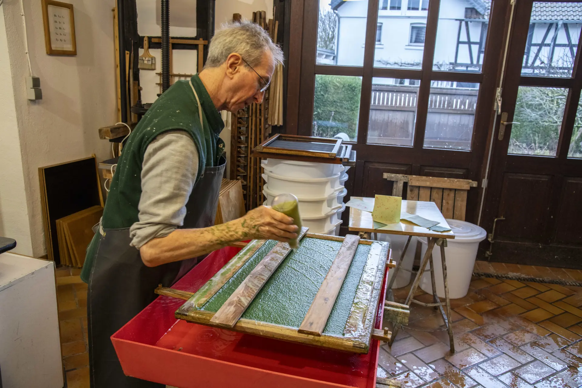 Ein Mann bei der Gestaltung eines frisch geschöpften Papierbogens mit der Technik des sogenannten „Pulp Paintings“ oder auf Deutsch „Papierguss“ – hier gießt er auf den grün geschöpften Bogen weißen Faserbrei.