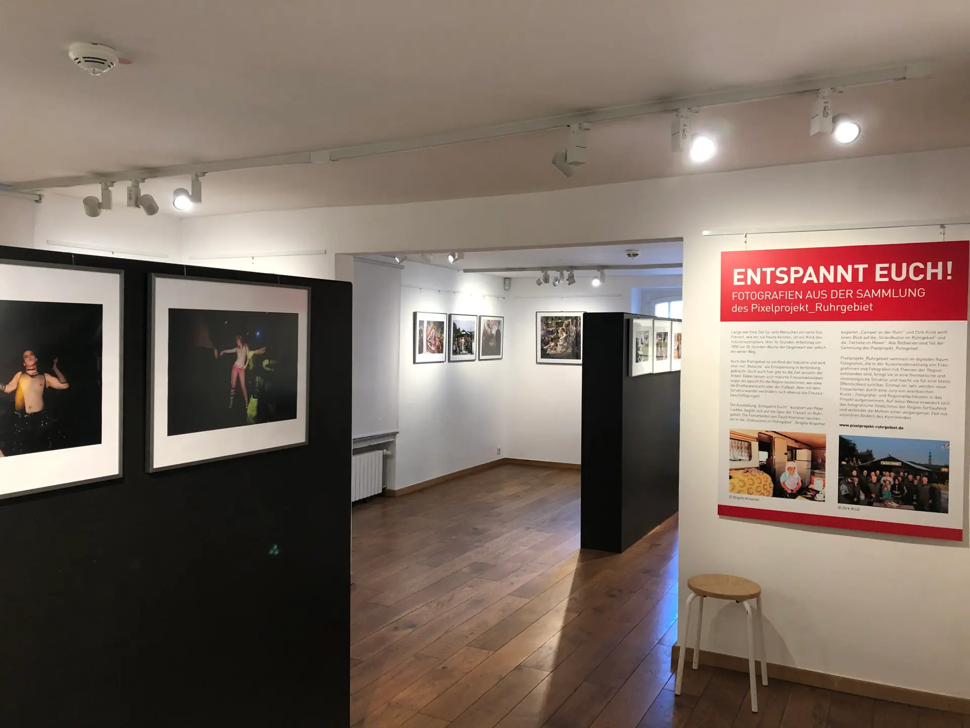 Blick in eine Ausstellung. An den Wänden hängen Fotos mit verschiedenen Motiven. An einer Wand hängt ein großes Plakat, auf dem "Entspannt Euch. Fotografien aus der Sammlung des Pixelprojekt_Ruhrgebiet".