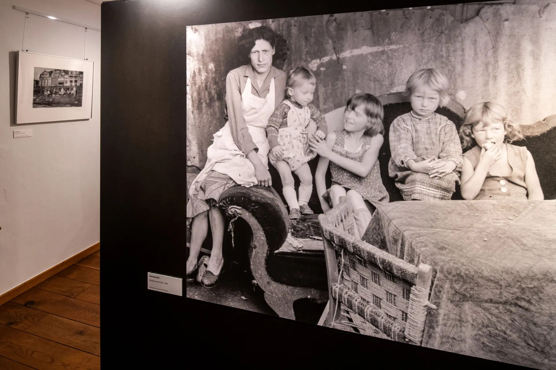 Blick in eine Ausstellung. Im Vordergrund ist eine große schwarz-weiss Fotografie. Auf dieser sitzt eine Frau mit vier Kindern auf einem Sofa. Vor ihnen steht ein Tisch.