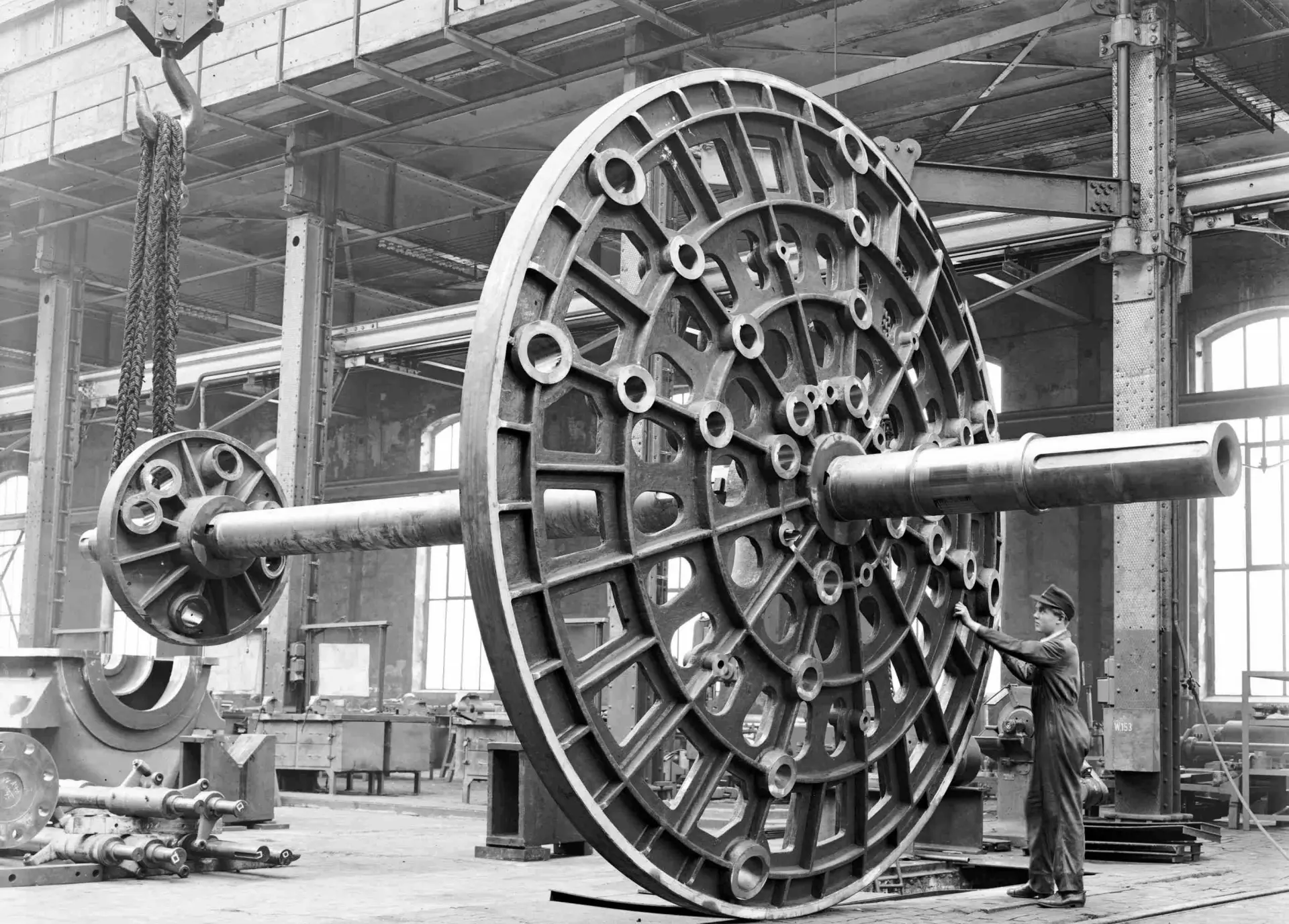 Schwarz-Weiß Fotografie einer großen Industrieanlage in einer Werkhalle. Im Vordergrund ist ein großes, rundes Metallbauteil mit vielen Öffnungen und Verstrebungen zu sehen. Links hängt eine Kette von einem Kran. Im Hintergrund stehen weitere Maschineteile und Gerüste. Rechts ist ein Arbeiter in Arbeitskleidung zu sehen.