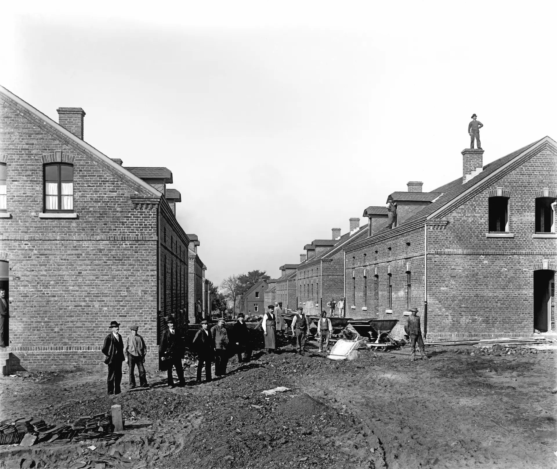 Historische schwarz-weiß Fotografie einer Siedlung. Zwischen den langen Backsteingebäuden arbeiten Männer. Im Vordergrund blicken einige Männer in dunklen Anzügen in die Kamera.
