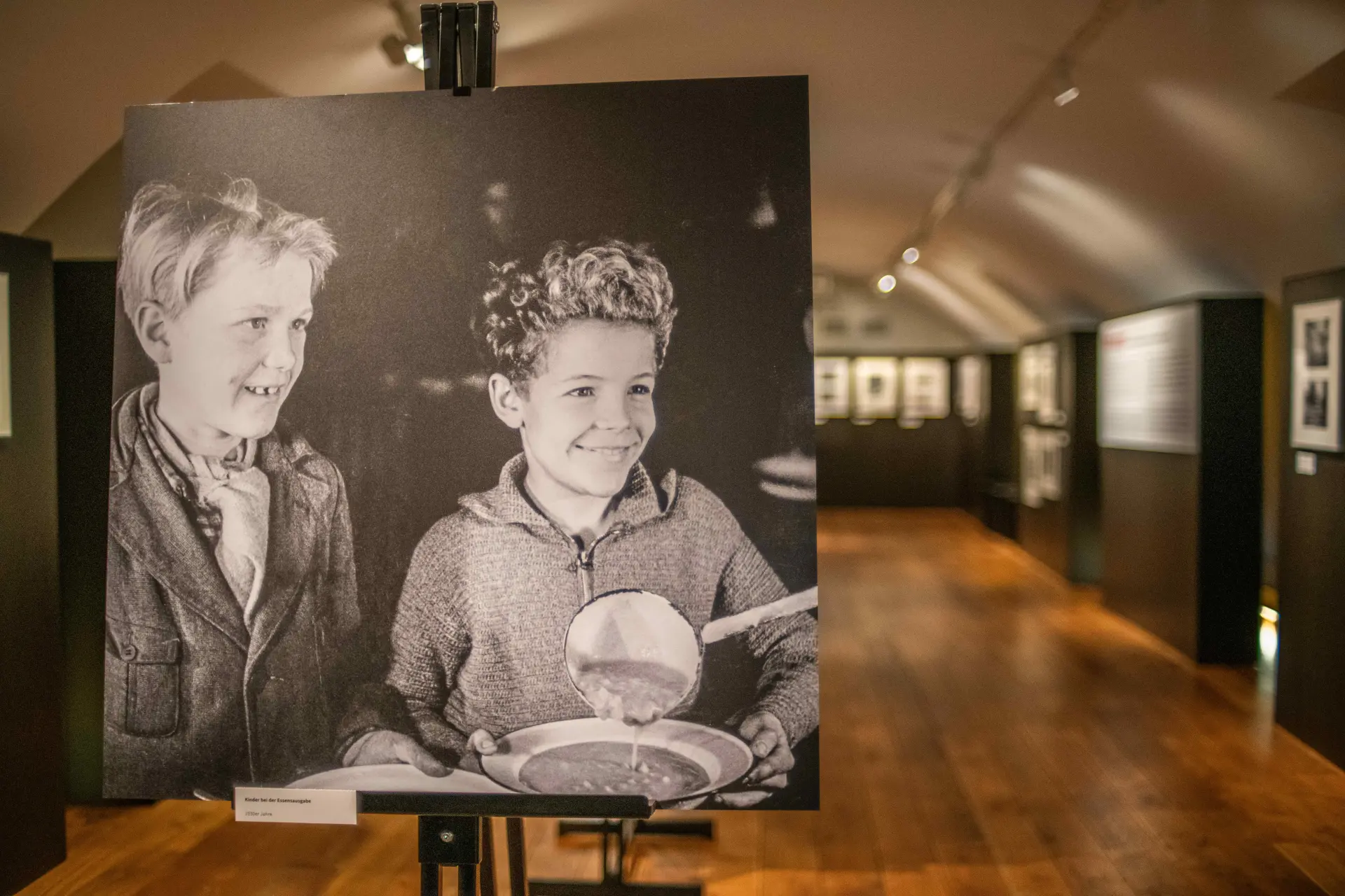 Blick in eine Foto-Ausstellung. Im Vordergrund ist eine historische schwarz-weiß Fotografie zu sehen, auf der zwei kleine Jungen Essen bekommen. Im Hintergrund sieht man weitere historische schwarz-weiß Fotografien.