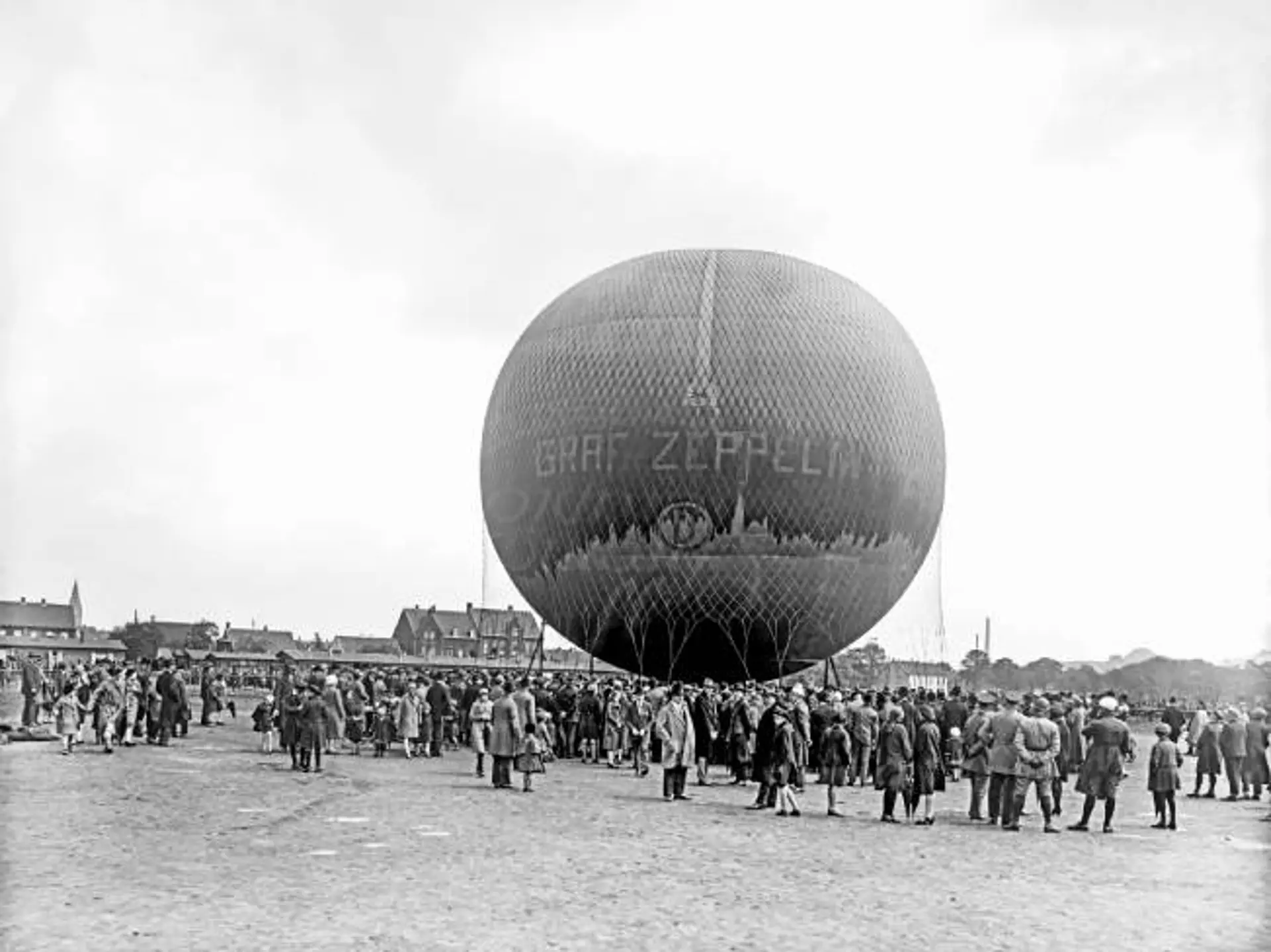 Historische schwarz-weiß Fotografie von einem Heißluftballon. Vor dem Heißluftballon steht eine große Menschenmenge, die den Ballon betrachtet.