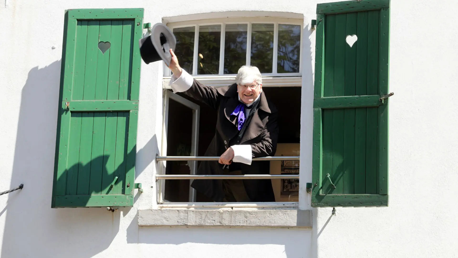 Ein Mann hebt seinen Zylinder zum Gruß an einem geöffneten Fenster