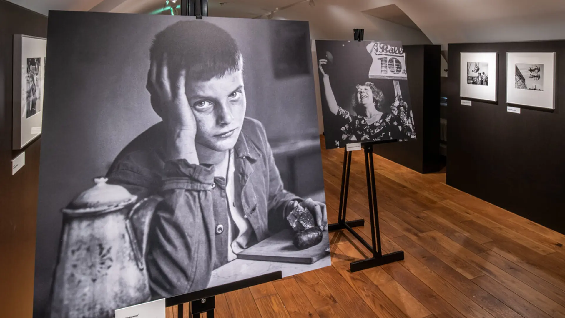 Blick in eine Ausstellung mit schwarz-weiß Fotografien. Im Fokus sind zwei auf Leinwänden aufgestellte Bilder mit einem traurig schauenden Jungen sowie einer Frau, die ein Schild mit Nummer hochhält.