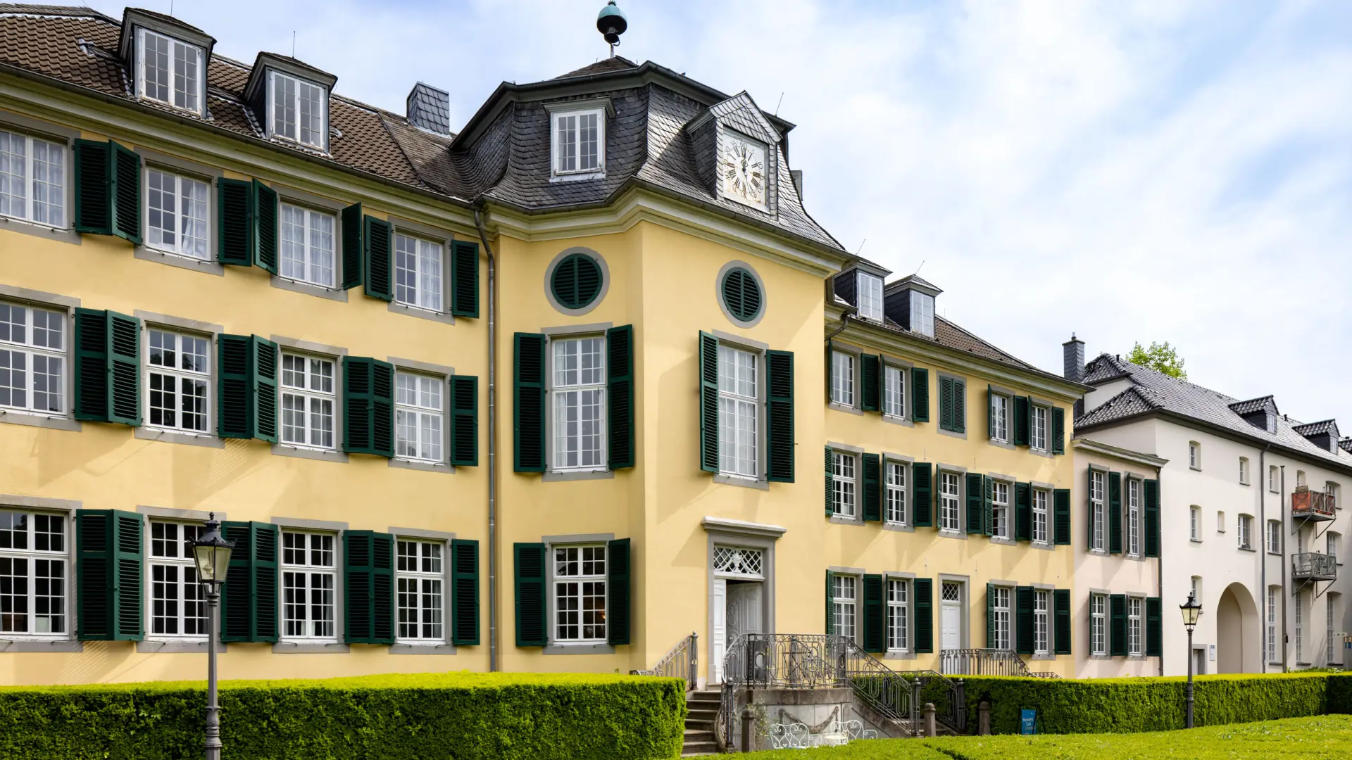 Steingebäude mit grünen Fensterläden. Davor steht eine Metallfigur, die an einen Mann im Anzug erinnert.