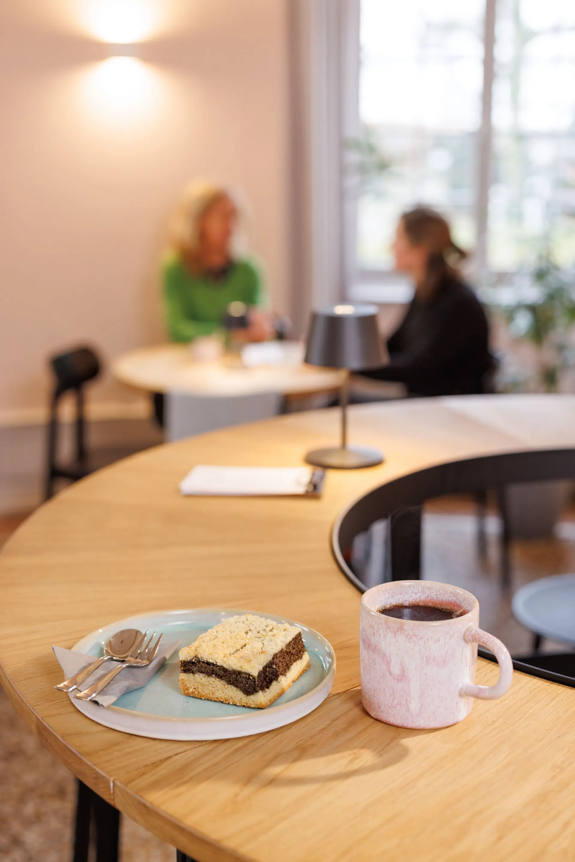 Im Café: Ein Tisch mit Kaffee und Kuchen, im Hintergrund sitzende Personen an einem Tisch