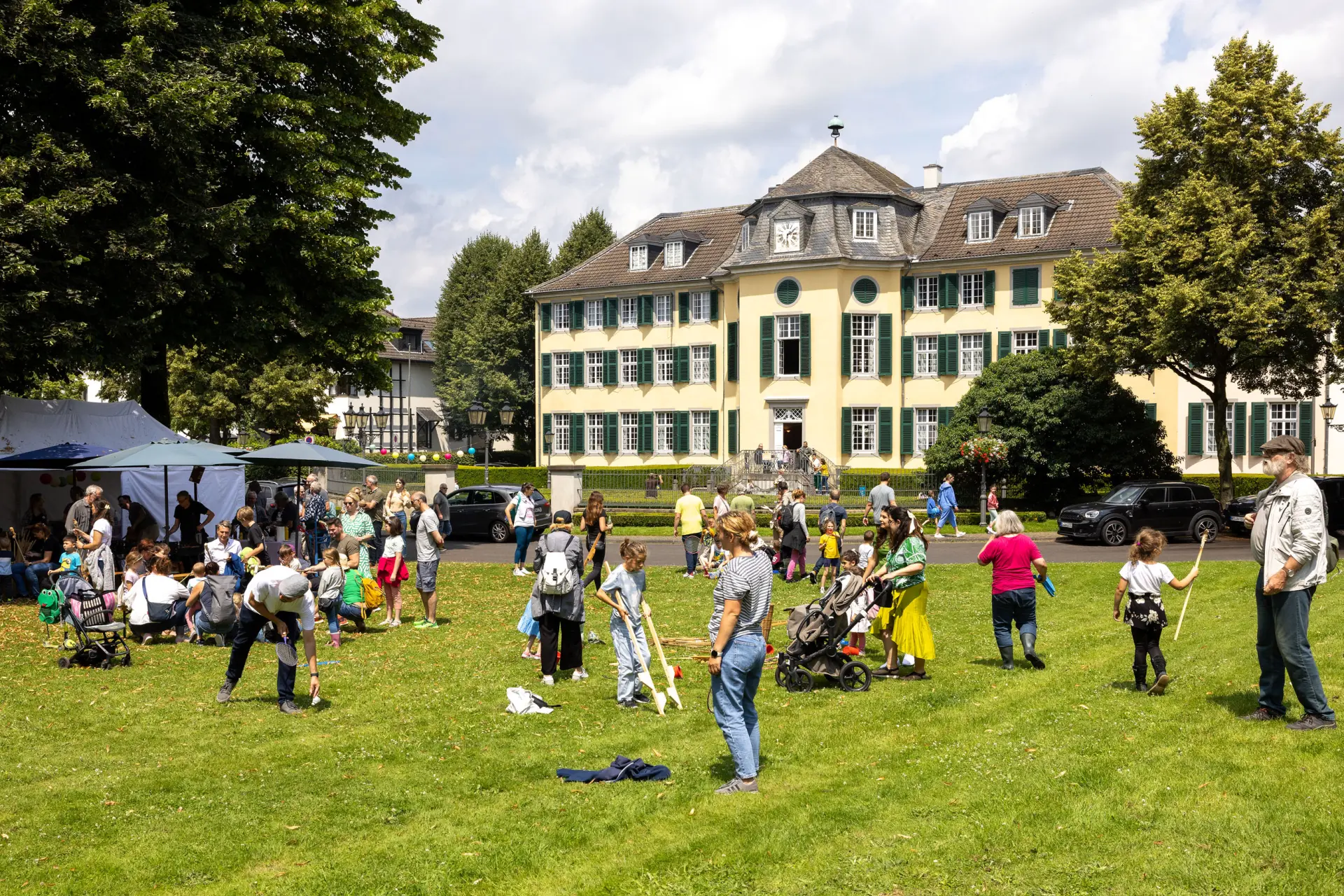 Viele Menschen auf einer Wiese. Im Hintergrund ein barockes Herrenhaus.