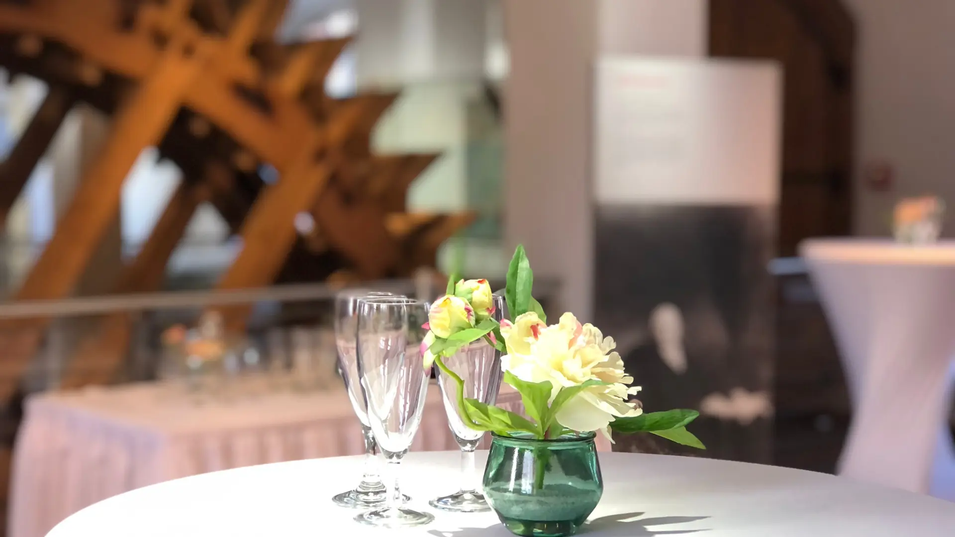 Ein Stehtisch mit weißer Tischdecke, einer Vase mit Blumen und Sektgläsern vor einem hölzernen Wasserrad