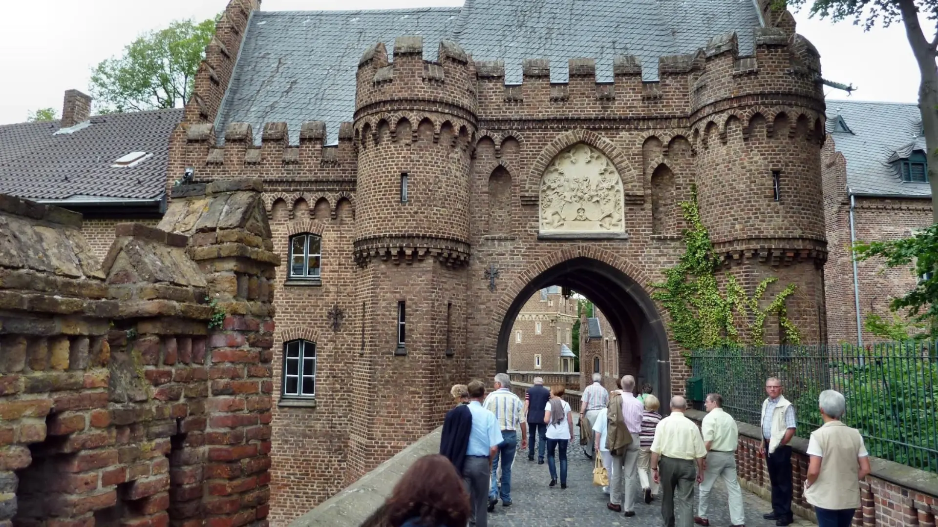 Eine Gruppe von Menschen läuft über eine Brücke auf eine Burg zu.