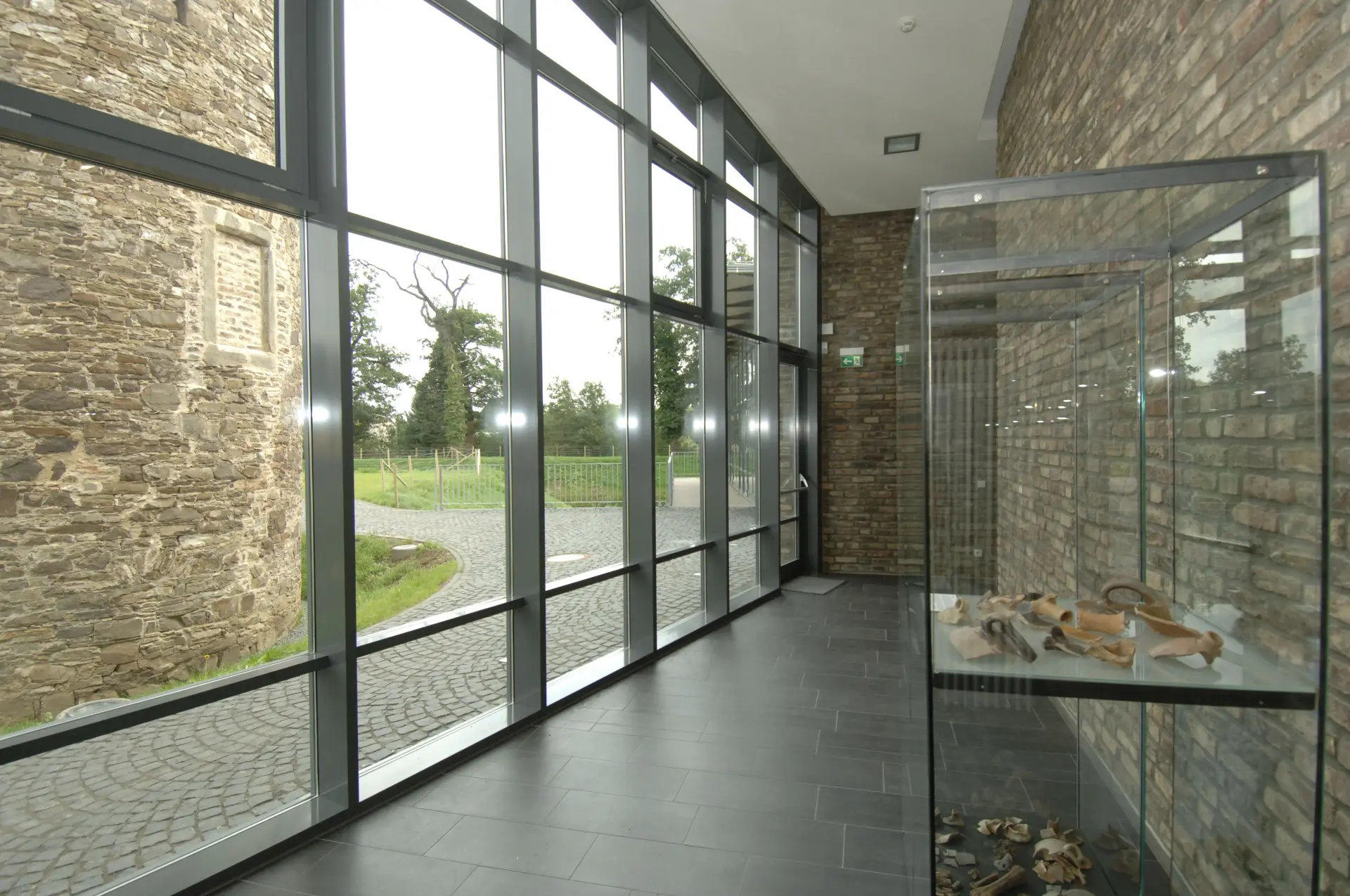 Ein Flur, der links eine Glasfront und rechts eine Steinwand hat. Darin steht eine Vitrine mit Tonscherben darin.