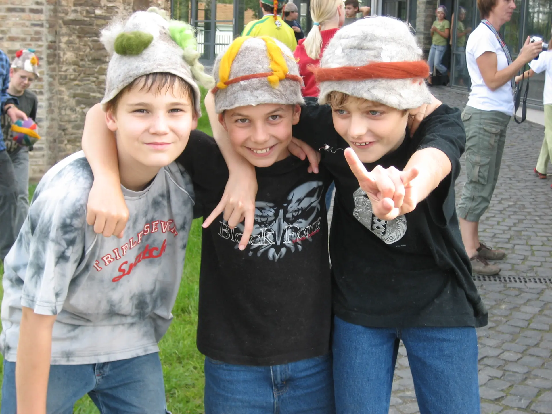 Drei Jungen mit Filzhüten liegen sich in den Armen und lachen in die Kamera.