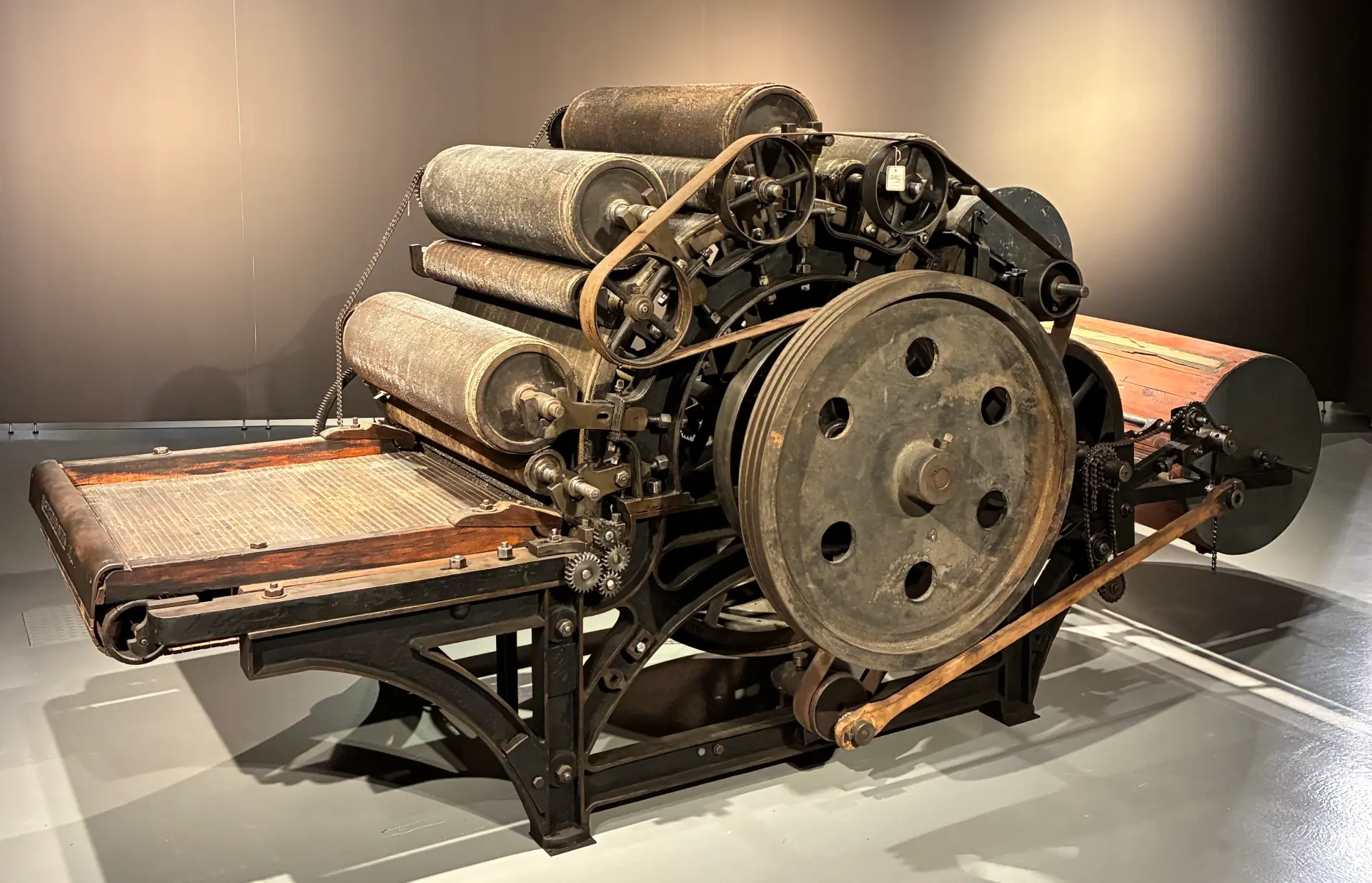 Große historische Textilmaschine aus Metall und Holz, ein sogenannter Krempel. Die Maschine steht frei im Raum eines Museums. Mehrere übereinander angeordnete Walzen sind oben auf der Maschine. Vorne befindet sich ein großes, rundes Schwungrad mit Löchern, das über Lederriemen und Zahnräder mit den Walzen verbunden ist. Unten trägt ein stabiles schwarzes Metallgestell die Konstruktion. Links ist eine flache Zuführfläche mit einem hölzernen Rahmen zu sehen, über die vermutlich Rohfasern in die Maschine eingezogen wurden.