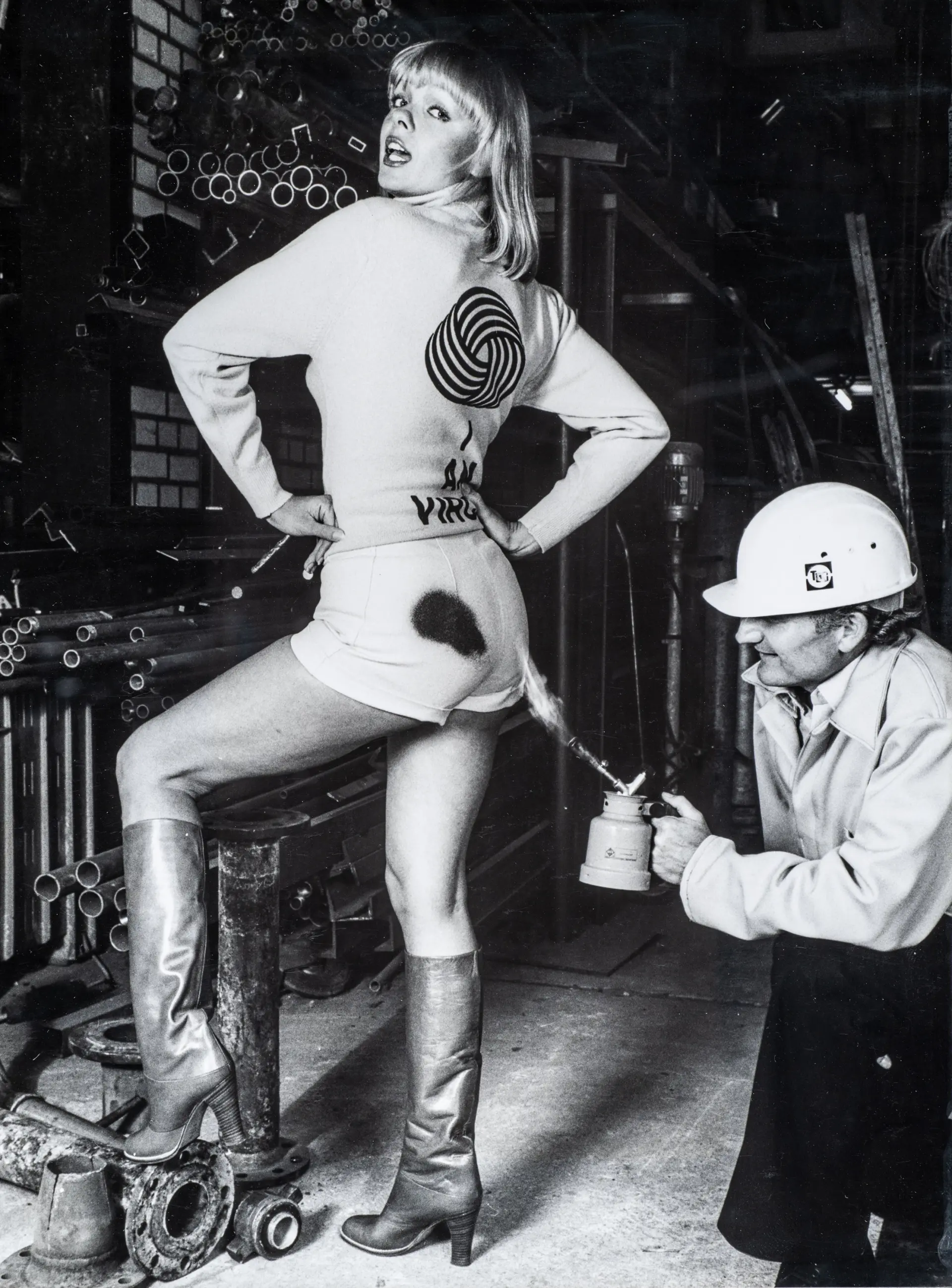 Schwarz-weißes Foto: Eine Frau in kurzen Shorts, langärmligem Oberteil und kniehohen Stiefeln steht in einer Werkhalle auf einem Metallteil und blickt über die Schulter zur Kamera. Auf ihrem Rücken ist ein großes Wollknäuel-Logo zu sehen. Ein Mann mit Schutzhelm kniet hinter ihr und richtet ein Gerät mit Flamme auf die Rückseite ihrer Shorts, wodurch ein dunkler Fleck entsteht. Im Hintergrund liegen Rohre und Metallteile, die die industrielle Umgebung betonen.