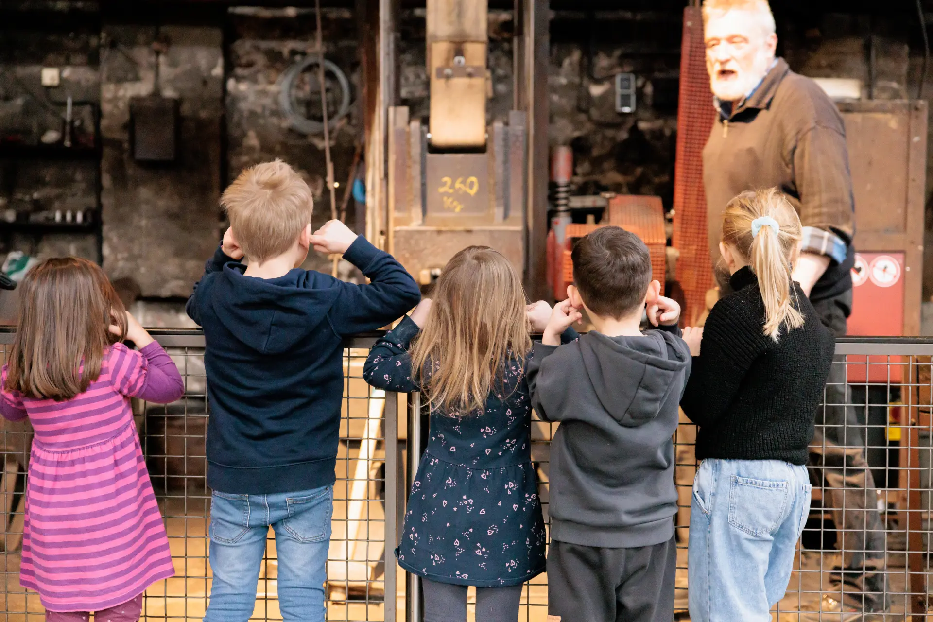 Eine Gruppe von Kindern von hinten, die sich die Ohren zuhält. Im Hintergrund ist ein Fallhammer zu erkennen.