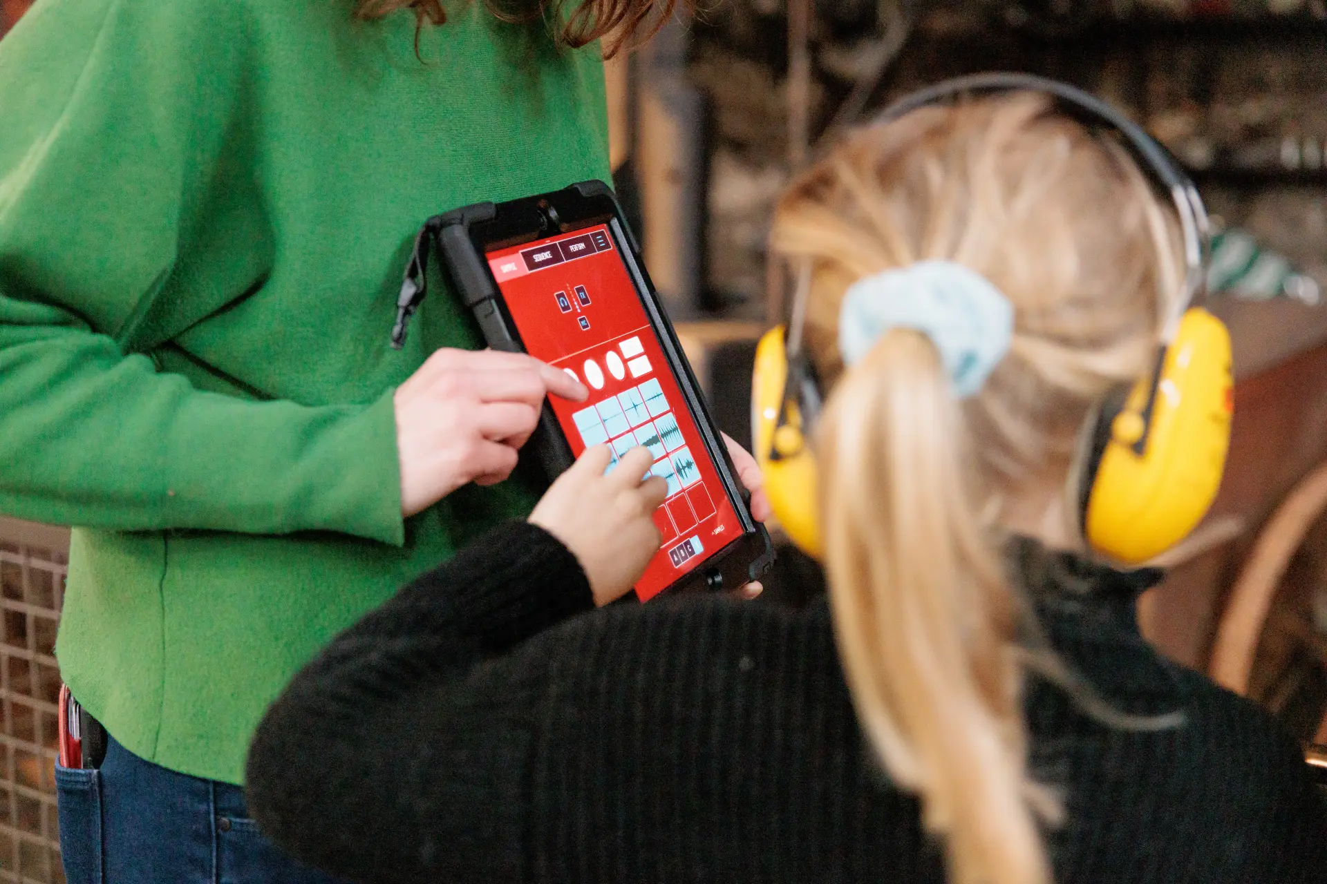 Ein Kind mit Ohrenschützern drückt auf ein Tablet, das von einer erwachsenen Person gehalten wird.