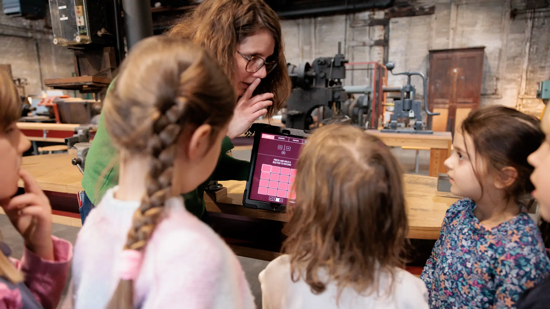 Einige Kinder stehen um eine Frau herum, die ein Tablet hält und durch einen Finger an den Lippen zeigt, dass alle leise sein müssen.