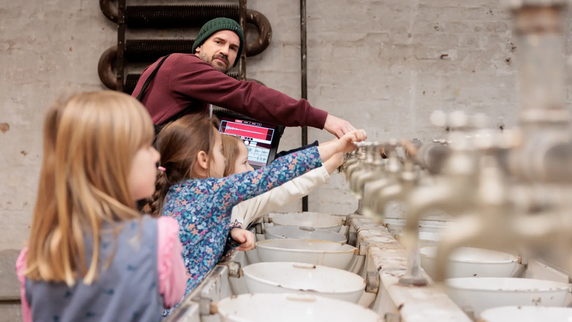 Ein Gästeführer steht mit einigen Kindern an alten Waschbecken und alle drehen an den Wasserhähnen.