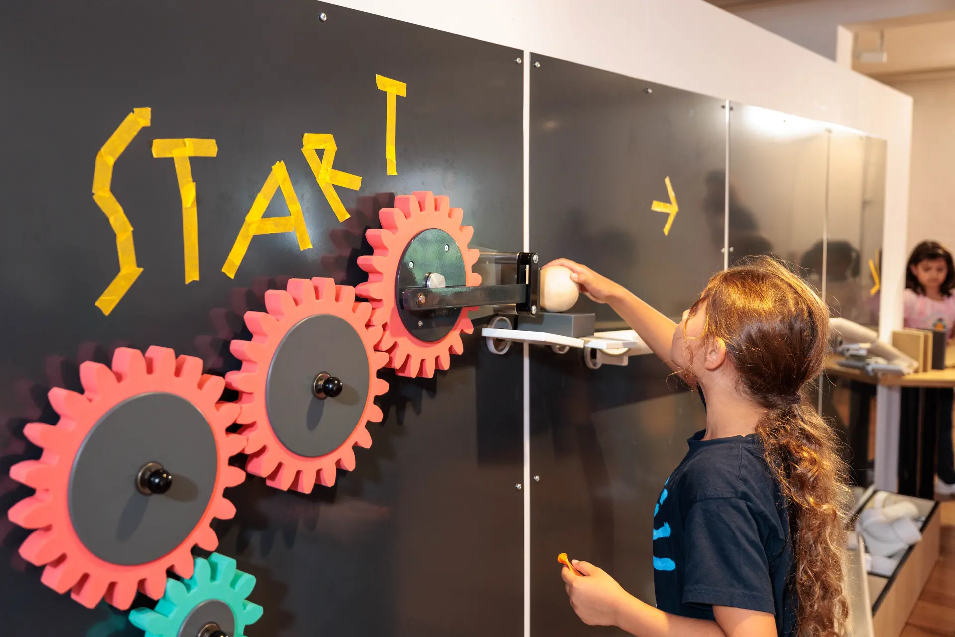 Ein Kind legt eine Kugel auf eine Bahn, die an einer Wand entlang führt. Vor der Bahn ist mit Tape das Wort "Start" geklebt.