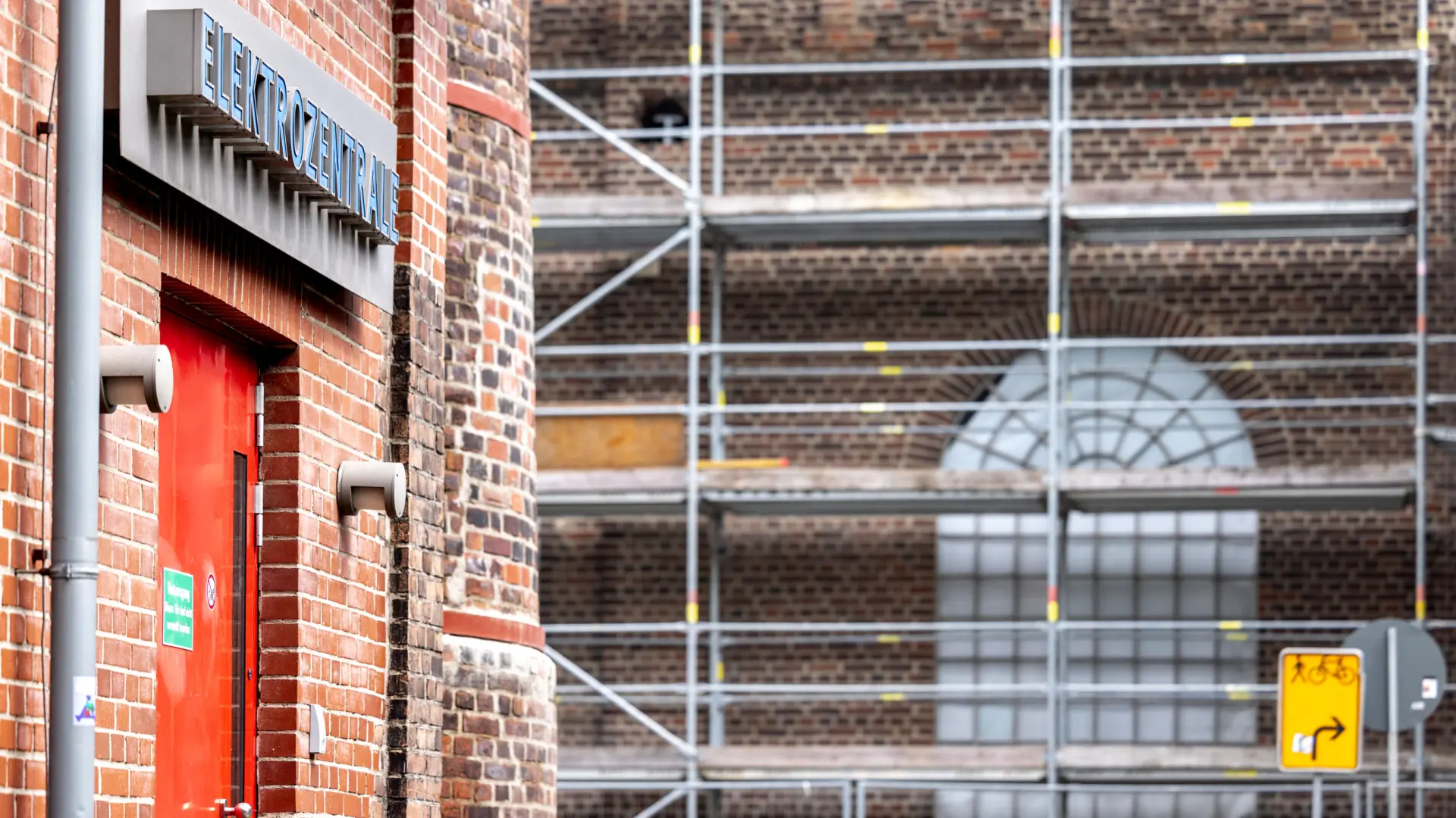 Ein Backsteingebäude links im Anschnitt, auf dem in blauen Buchstaben "Elektrozentrale" steht. Im Hintergrund steht ein Baugerüst vor einer Backsteinwand mit großem Fenster.