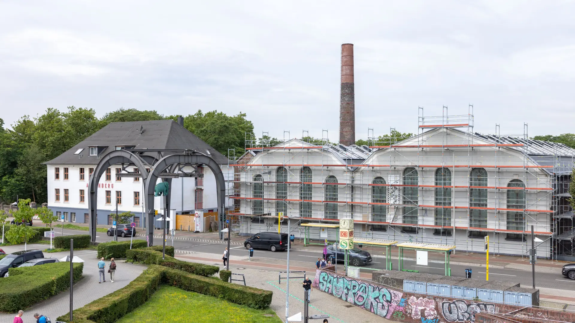 Blick über einen Platz und eine Straße hinaus auf zwei weiße Gebäude. Eines der Gebäude ist von einem Baustellengerüst umgeben.