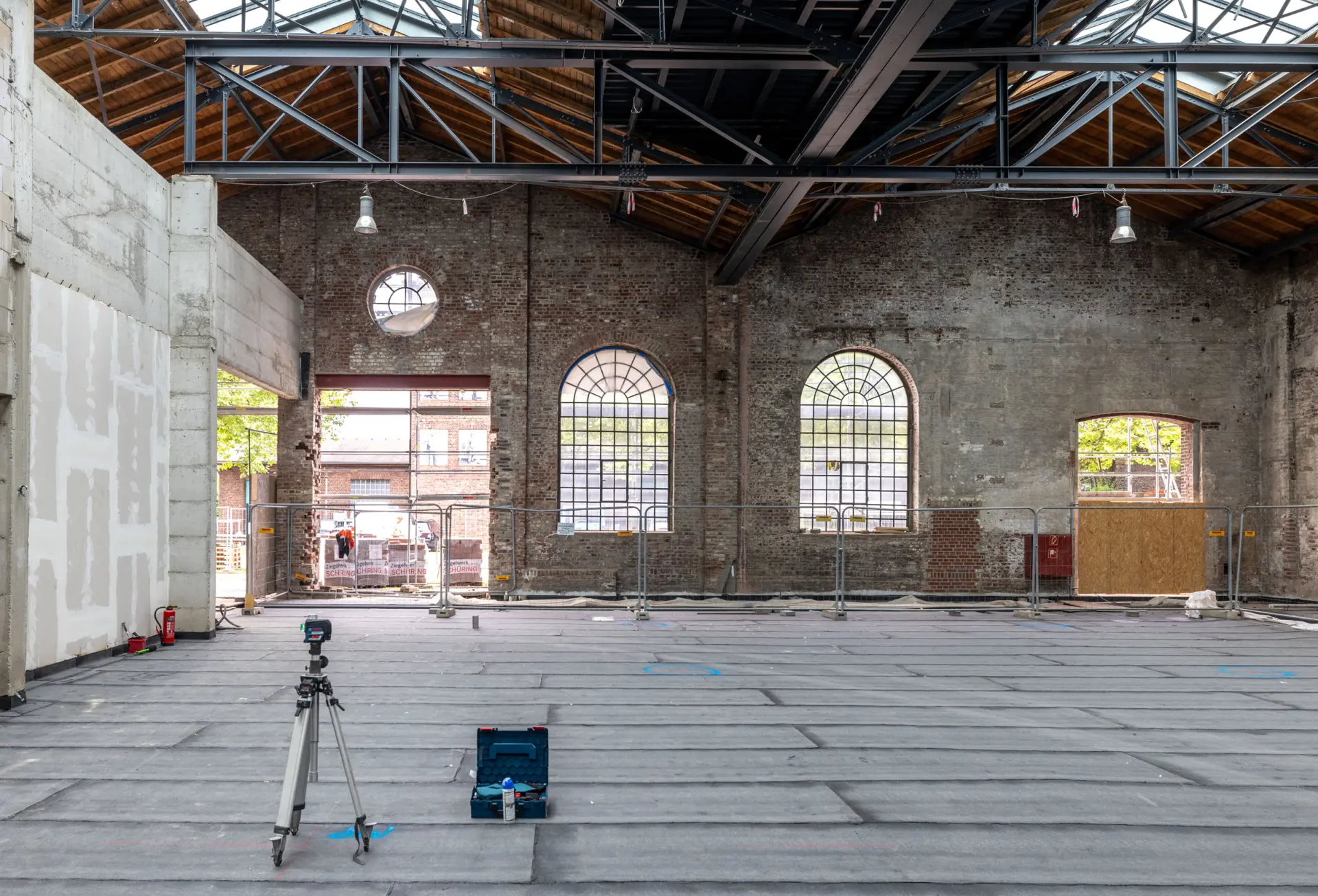 Eine leere Halle, in der Bauarbeiten stattfinden. Zäune begrenzen den Durchgang vor einer Wand mit großen Fenstern und einer großen Öffnung.