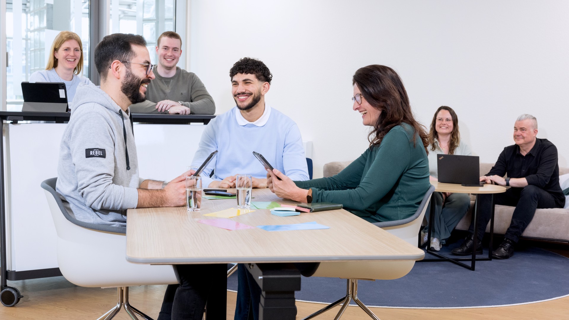 Im Vordergrund sind drei Personen an einem Tisch, im Hintergrund sitzen zwei Gruppen mit jeweils zwei Personen zusammen