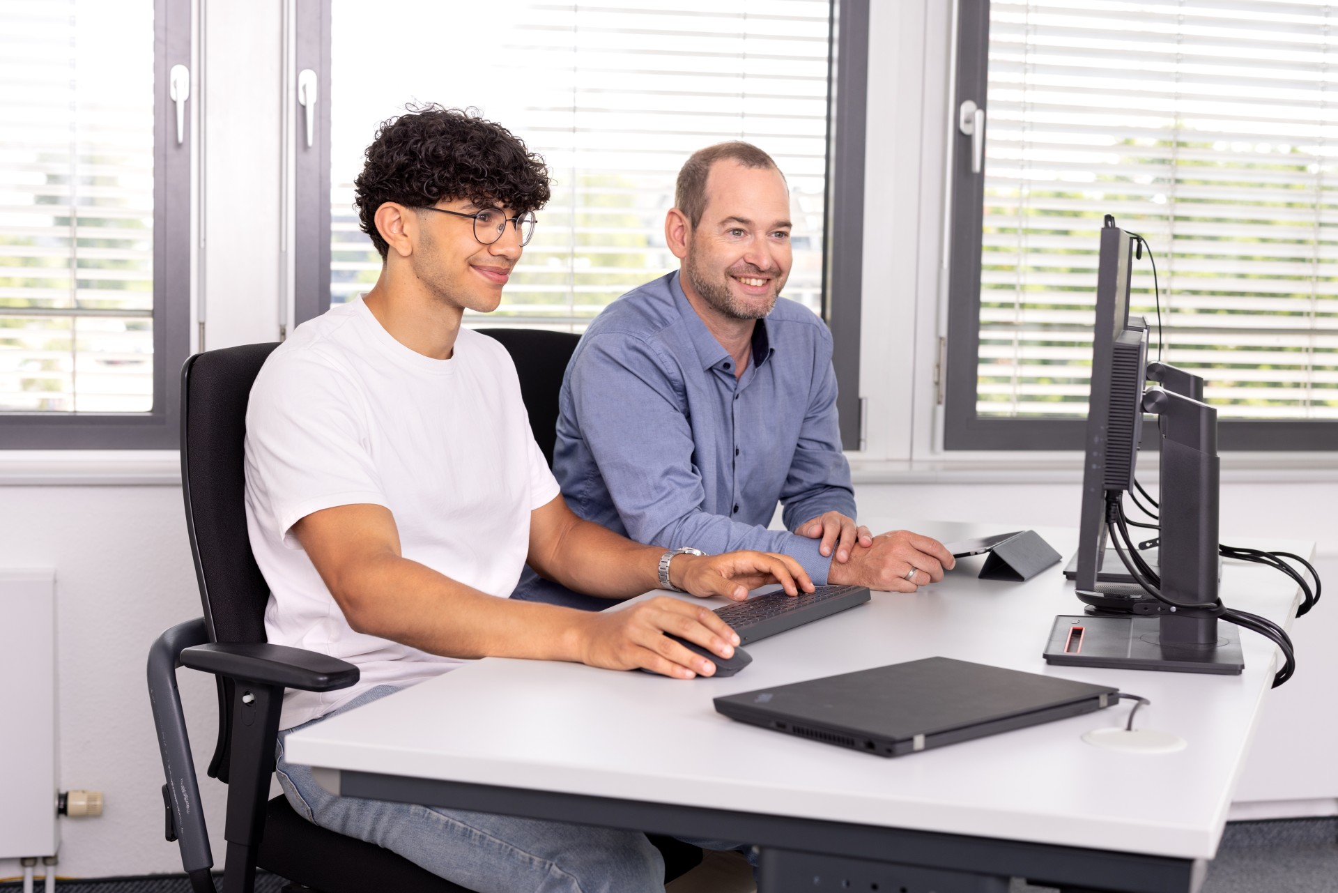 Eine Nachwuchskraft und ein Ausbilder sitzen an einem Schreibtisch und schauen auf einen Monitor