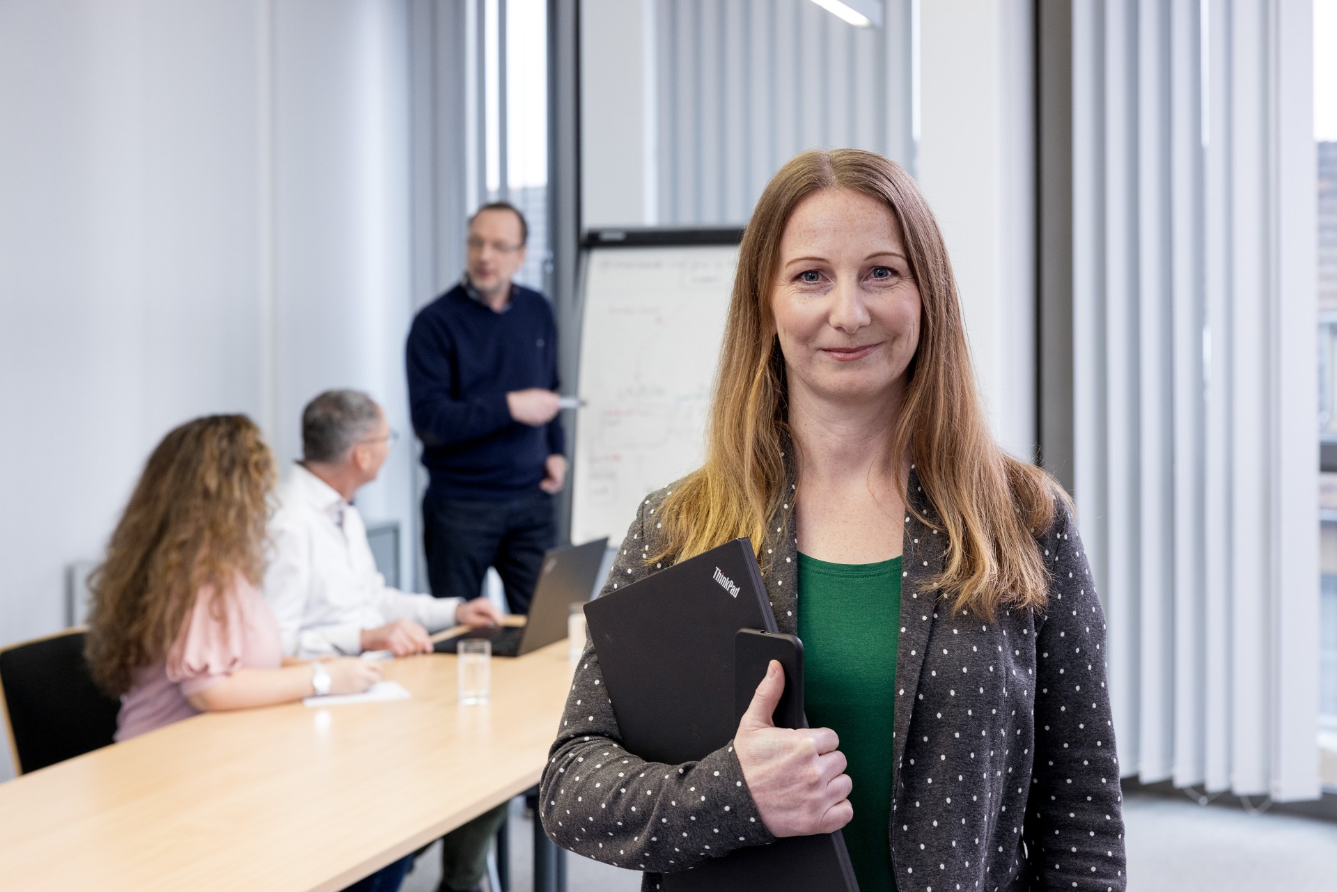 Eine Frau mit Laptop steht im Vordergrund, im Hintergrund sind 2 Frauen und ein Mann in einer Gesprächssituation zu sehen.