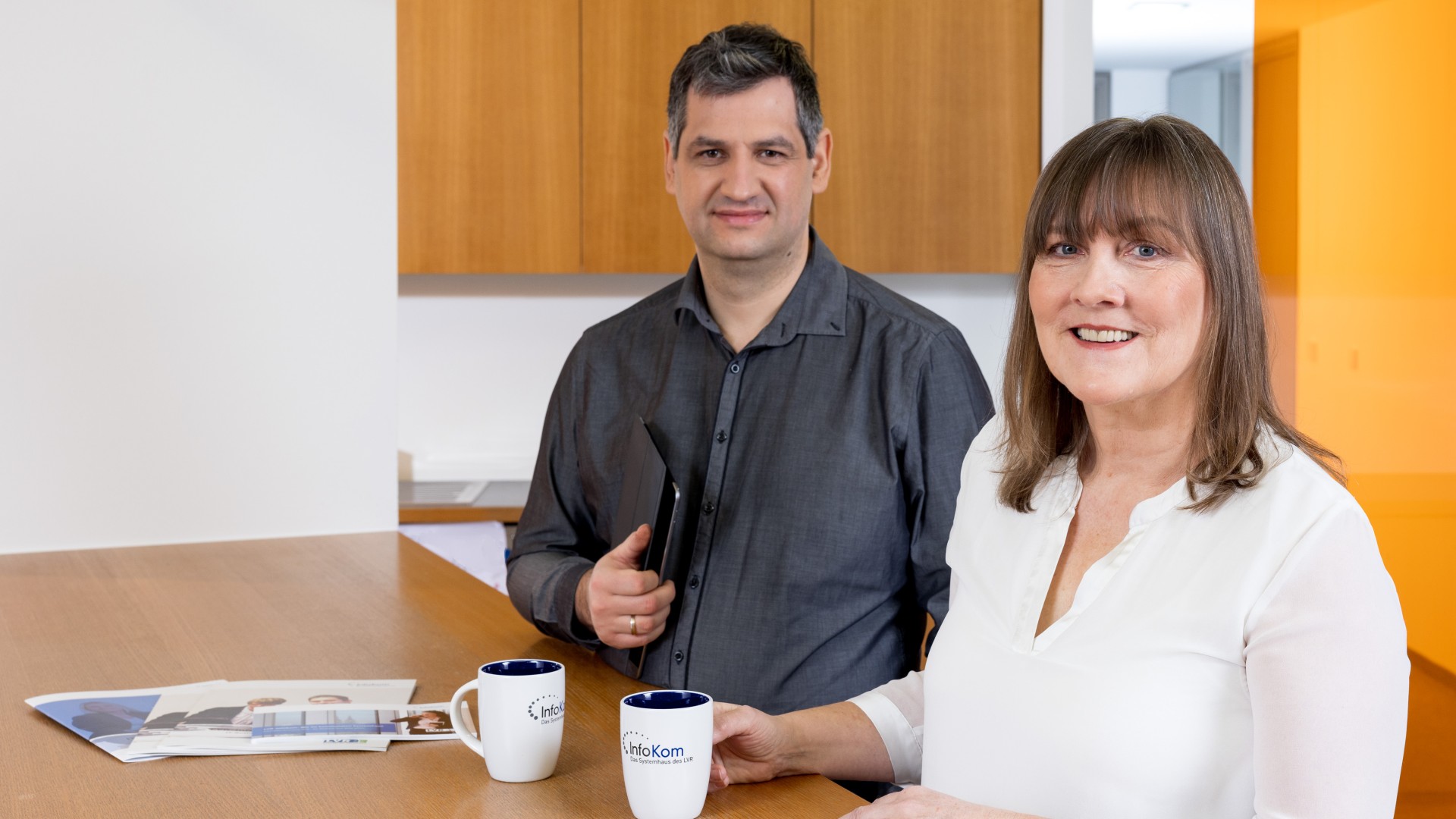 Zwei Männer sitzen an einem Tisch mit einem Magazin und einem Tablet. Zwischen ihnen steht eine lächelnde Frau und deutet auf das Magazin.