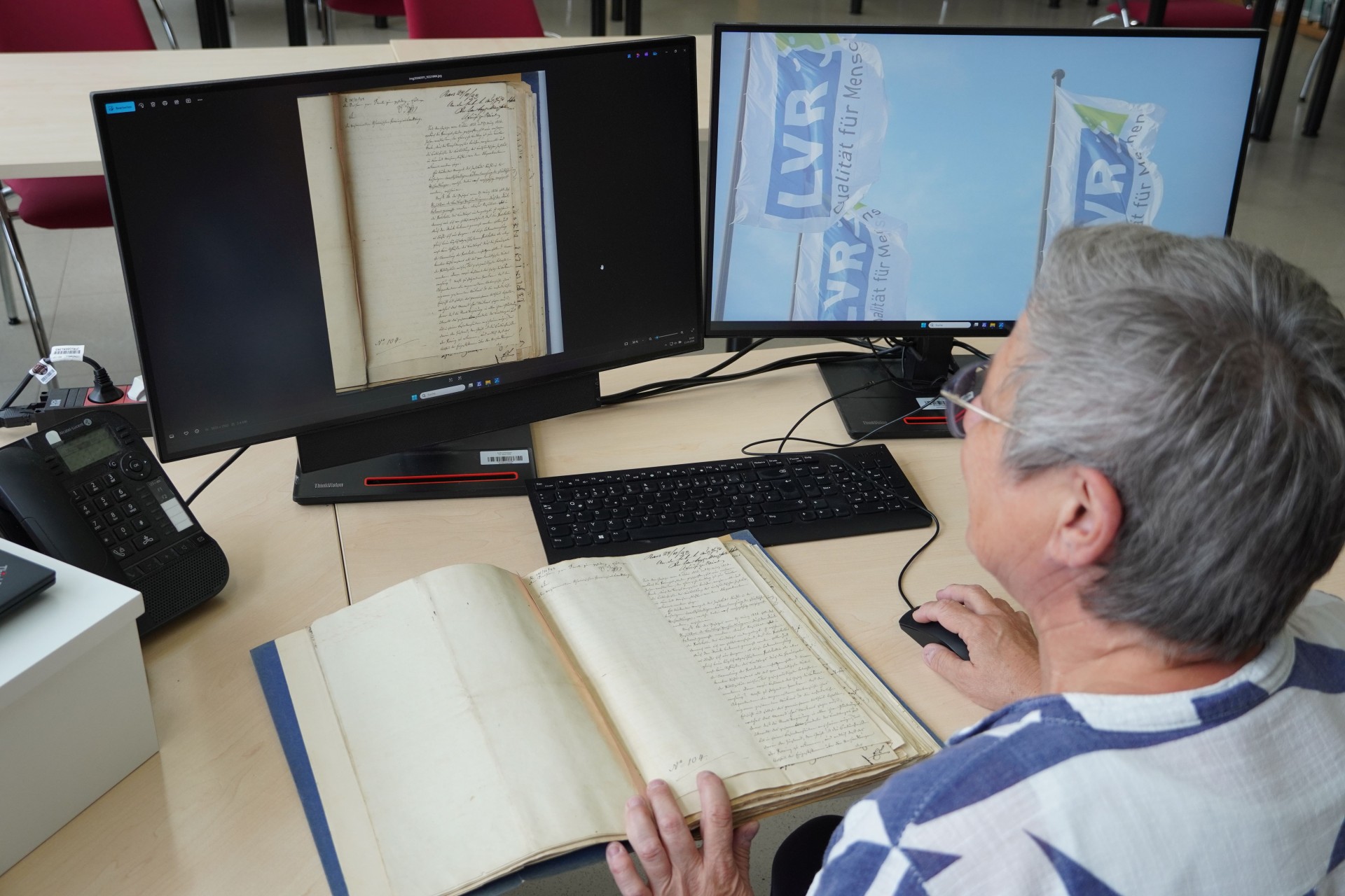 Eine Frau sitzt an einem Schreibtisch vor zwei Monitoren. Auf dem Tisch liegt ein Digitalisat.