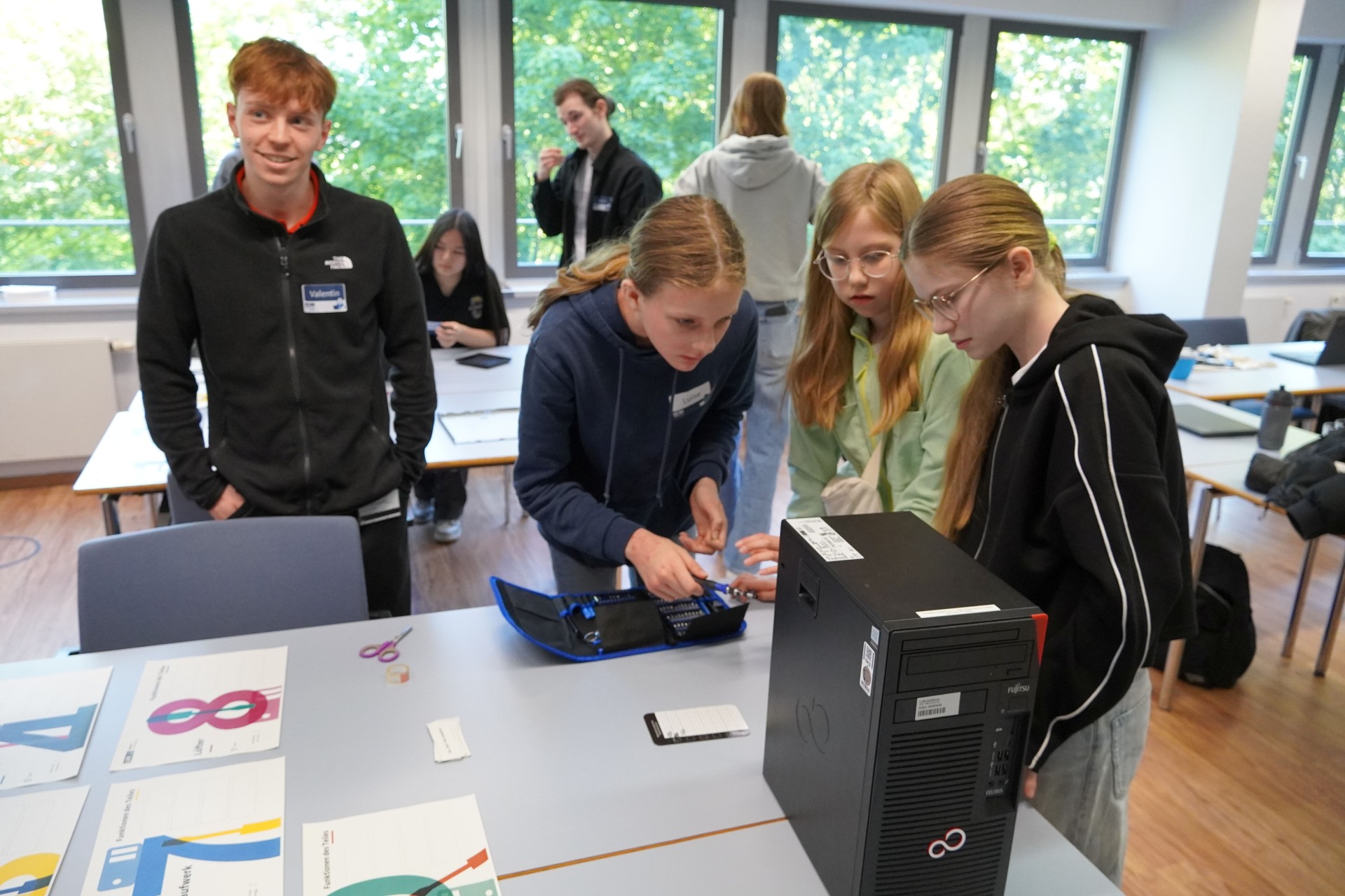 Drei Mädchen basteln an einem Computer, der auf einem Tisch steht. Daneben steht ein junger Mann.