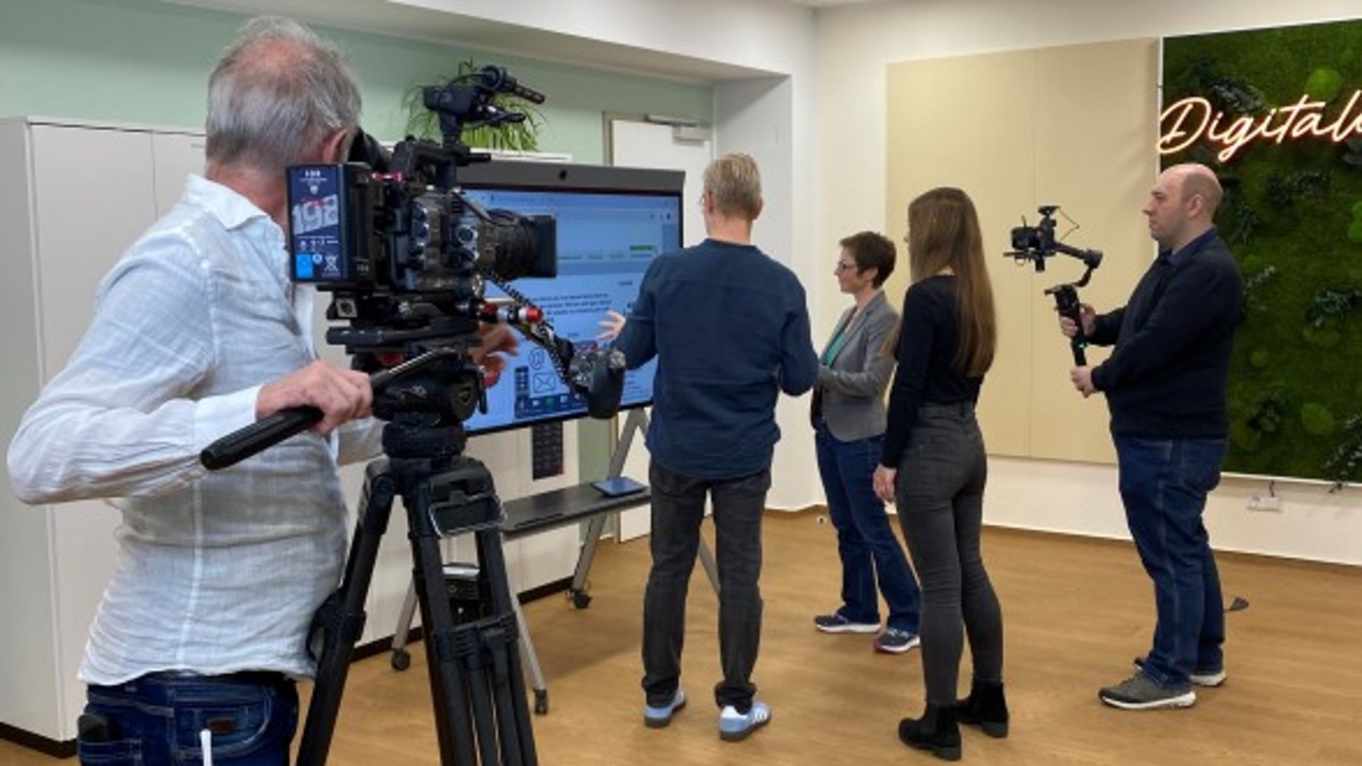 Menschen stehen vor einem Bildschirm, zwei Kameraleute, filmen sie dabei