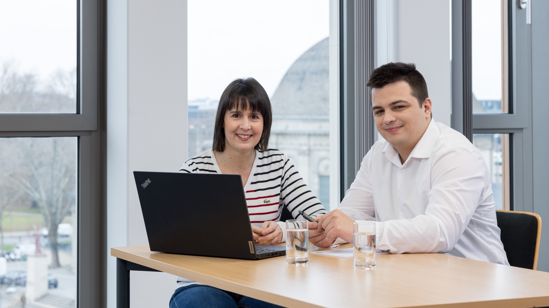 Eine Frau und ein Mann sitzen an einem Schreibtisch vor einem Laptop und lächeln in die Kamera.
