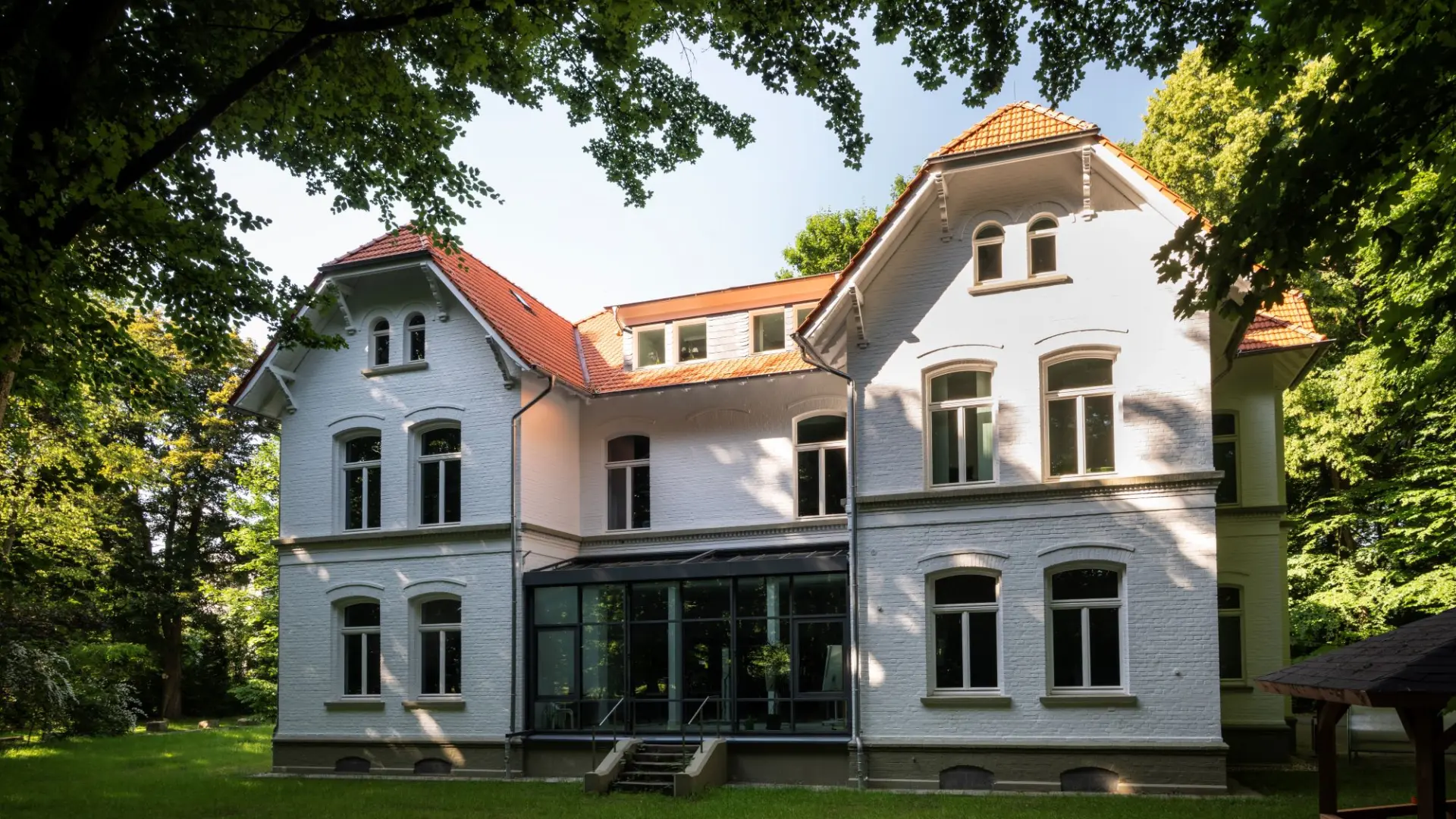 Historisches, weißes Backsteingebäude der Bildungsakademie mit rotem Ziegeldach, umgeben von Bäumen und einer Rasenfläche.