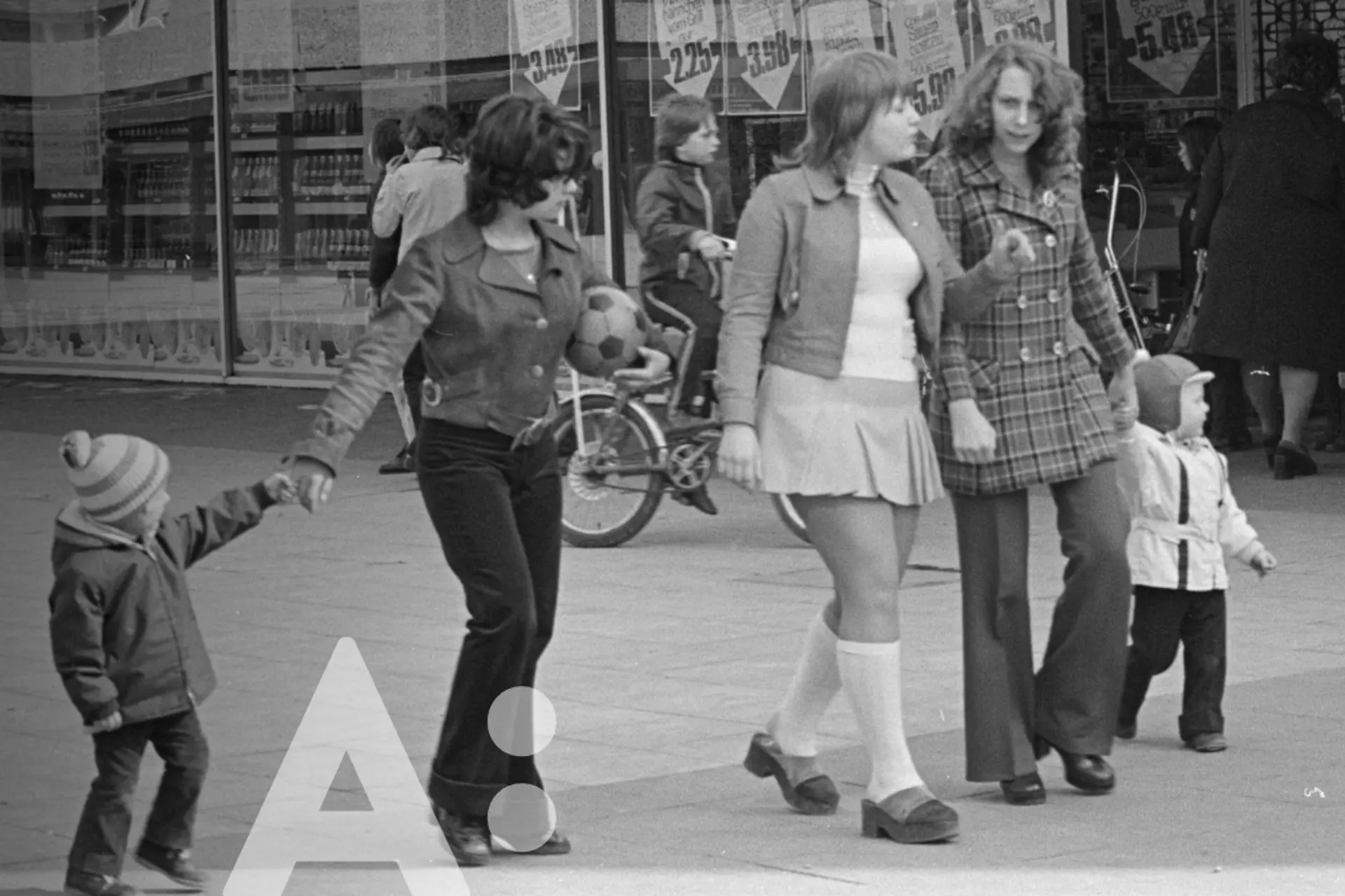 Fünf Personen gehen gemeinsam eine Straße entlang. In der Mitte gehen drei Frauen, flankiert von zwei Kleinkindern, jeweils einem auf der linken und rechten Seite.