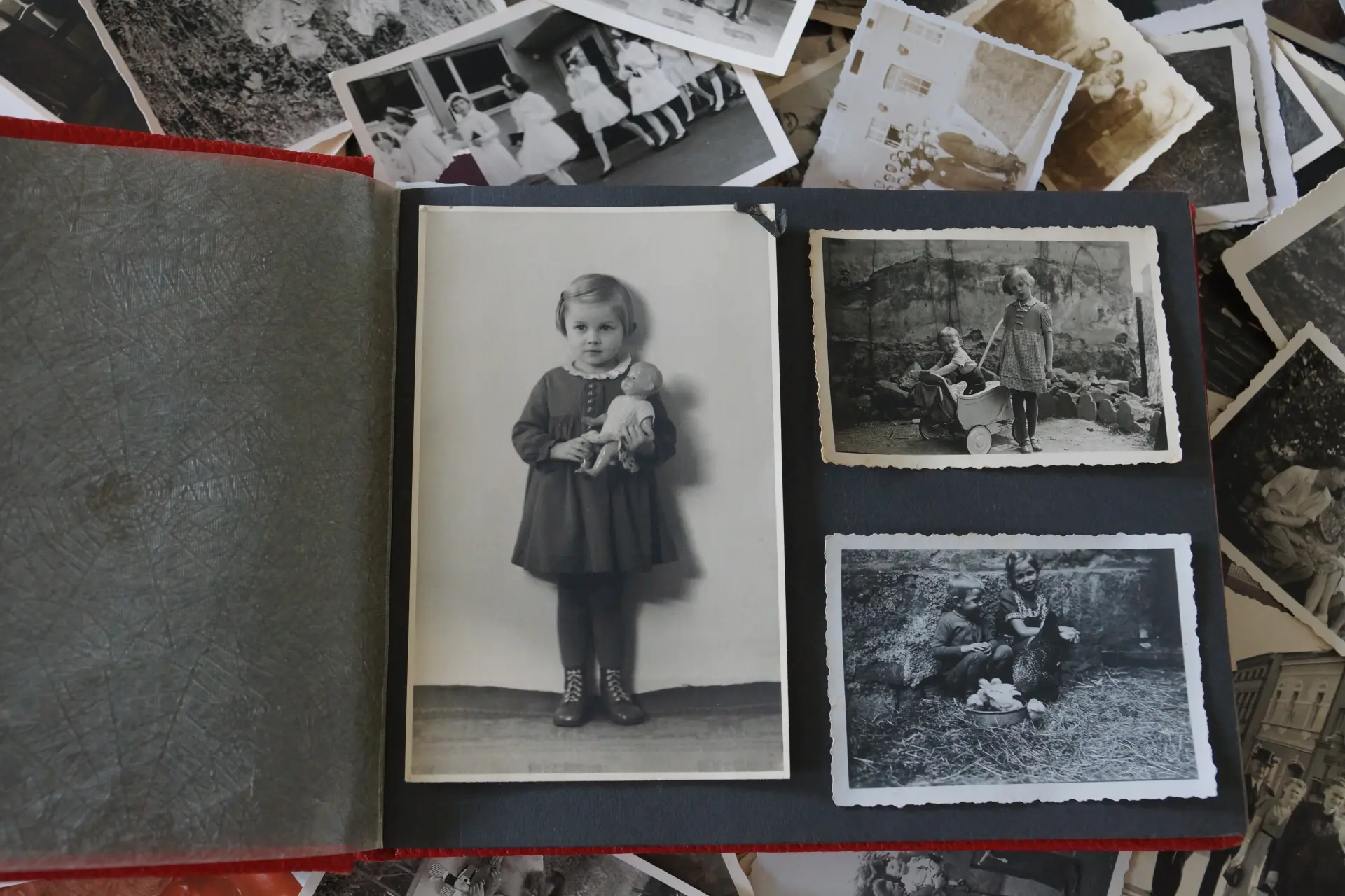 Aufgeschlagenes Fotoalbum mit rotem Einband und schwarzen Seiten. Auf der rechten Seite des Albums sieht man drei schwarz-weiß Fotos. Auf dem Linke im Hochformat ist ein Mädchen in Kleid mit weiße, Kragen abgebildet, das eine Puppe im Arm hält.