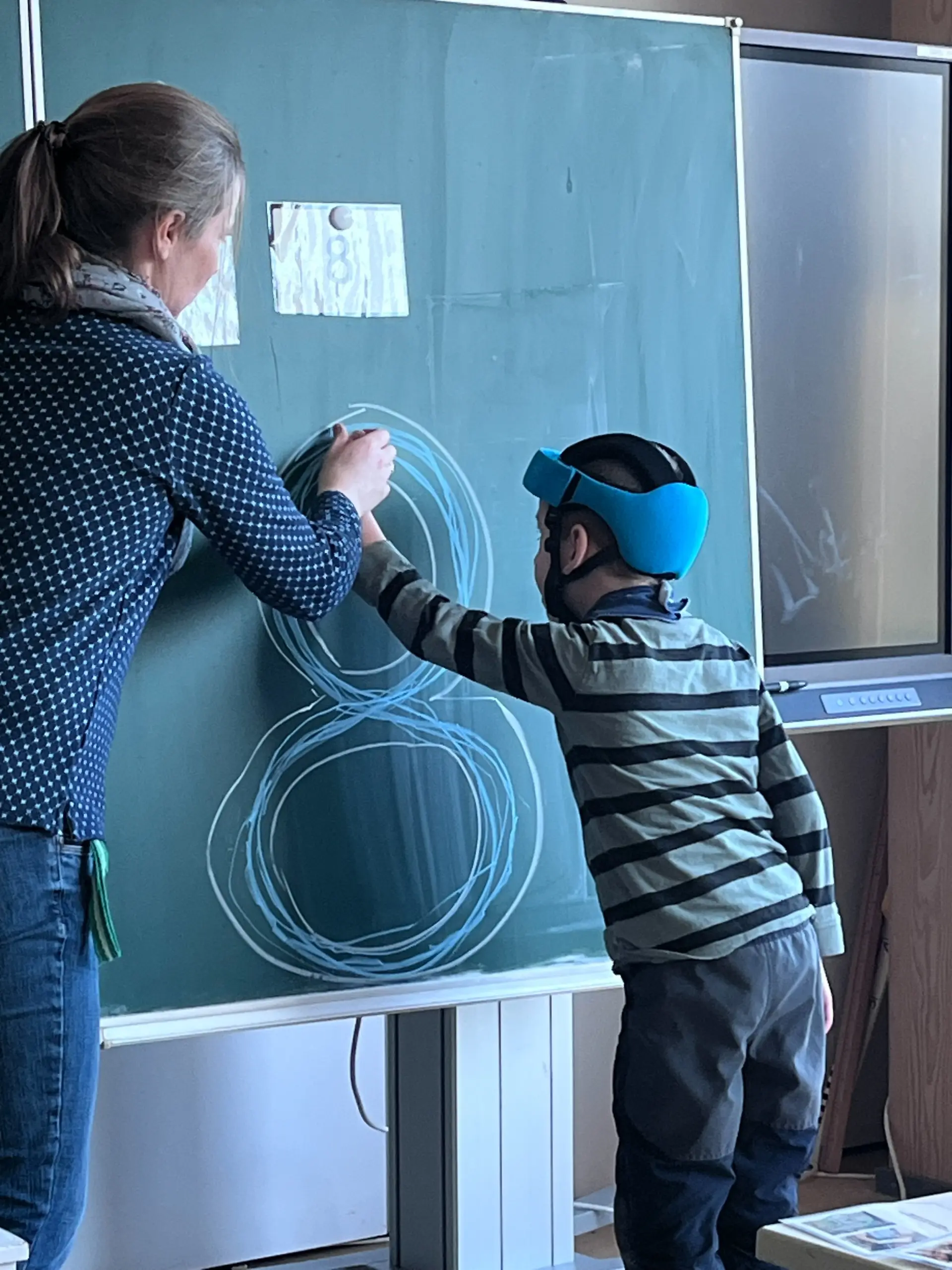 Primarstufe: ein Kind schreibt die Zahl "8" an die Tafel, eine Lehrkraft hilft