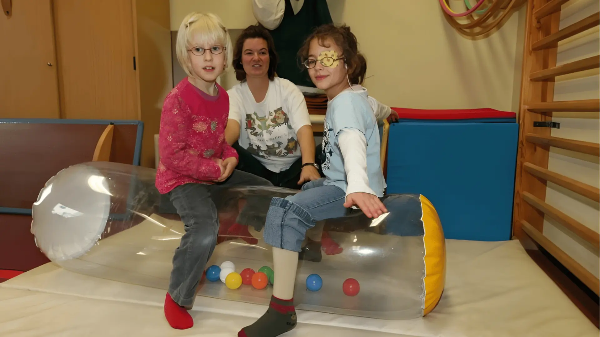 Zwei Kinder sitzen auf einer aufblasbaren Sitzeinheit und machen Physiotherapie mit einer Therapeutin