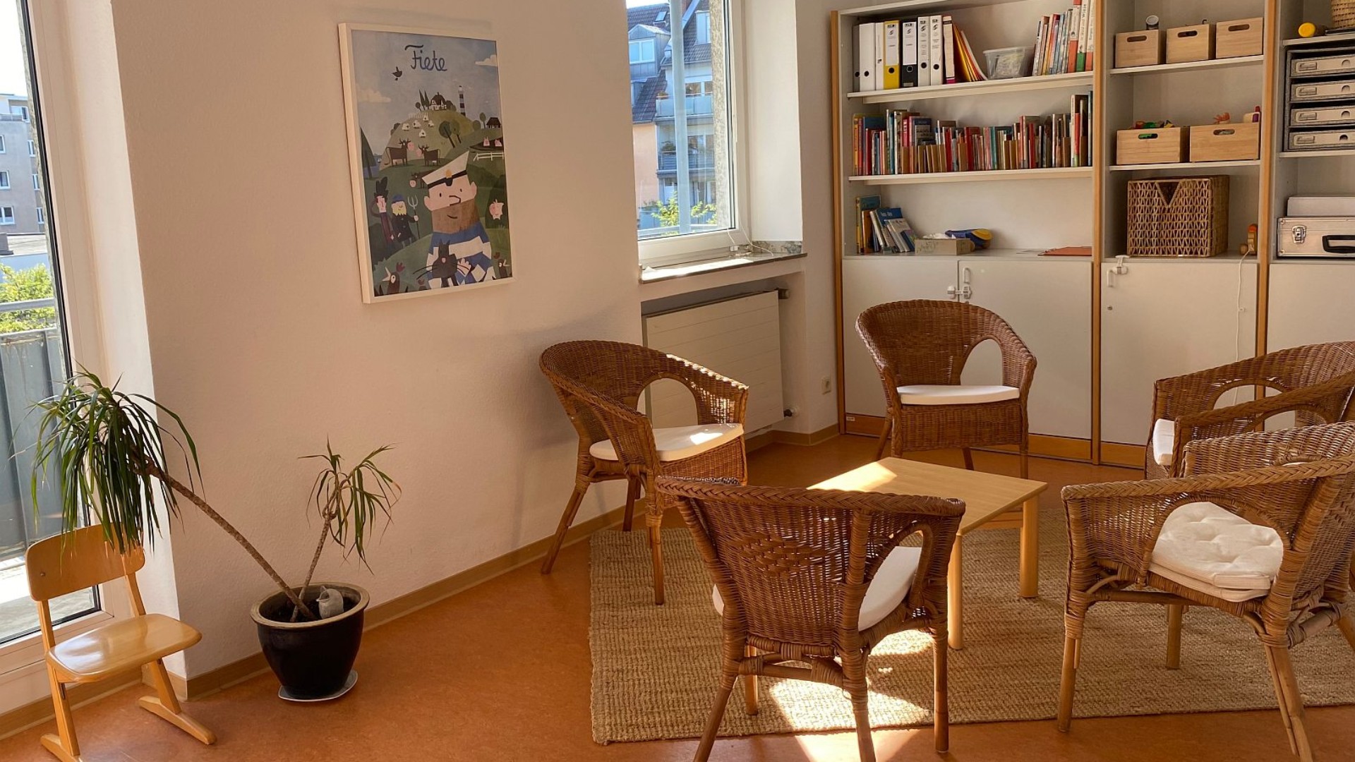 Blick in den Beratungsraum der Beratungsstelle: ein kleiner Tisch mit fünf Korbsesseln. Hinten an der Wand ein Regal mit Büchern und Schubkästen.