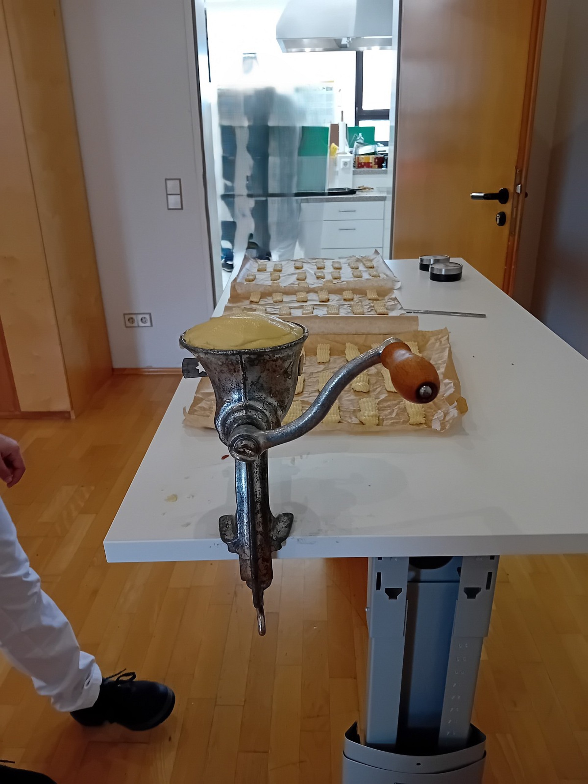 An einem Tisch ist ein Fleischwolf aus Gußeisen mit Teig befüllt. Auf dem Tisch stehen drei Bleche mit Spritzgebäck.