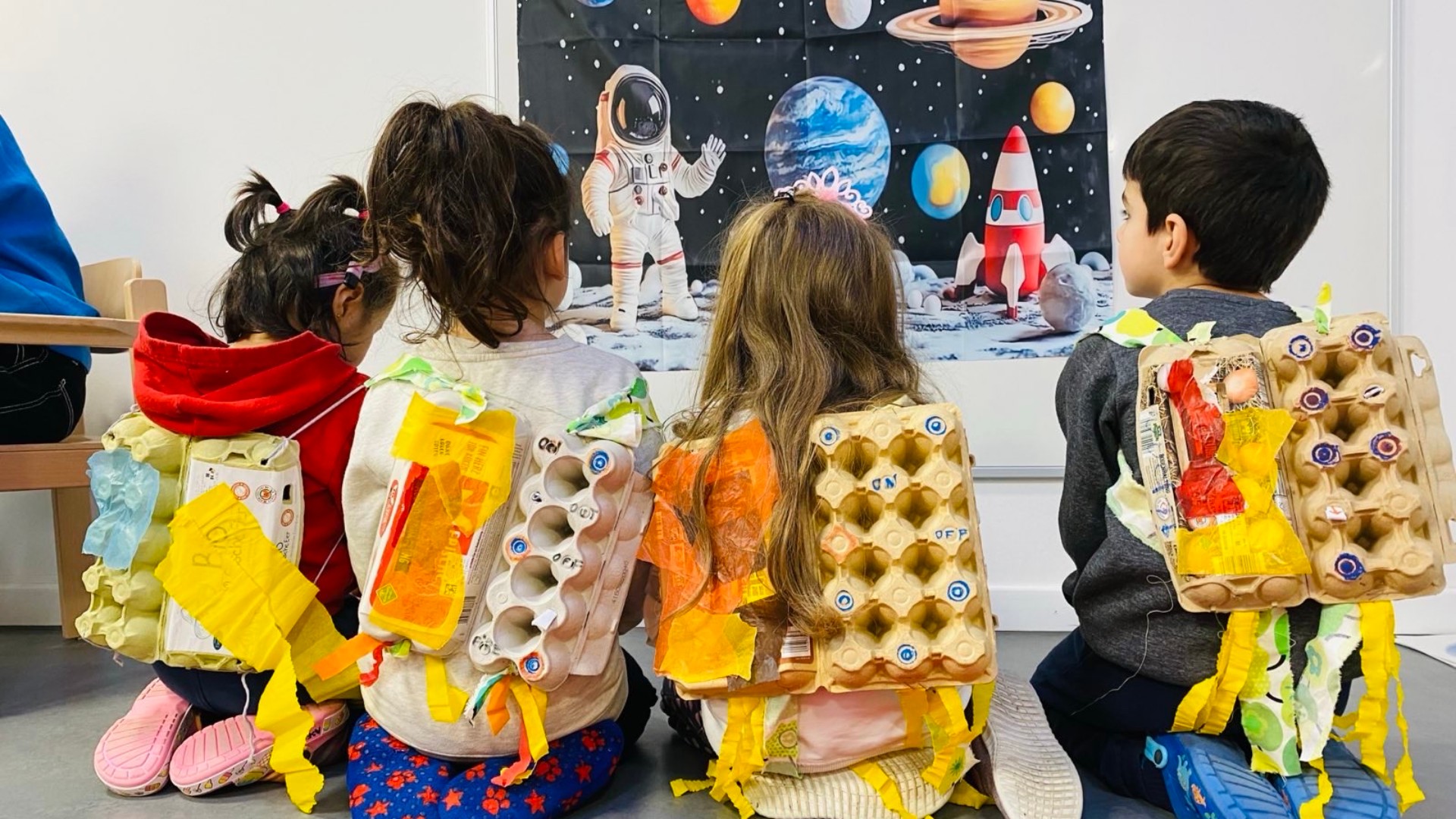 Kinder von hinten vor einem Planetenposter. Auf dem Rücken haben sie selbstgebastelte Antriebsraketen aus Eierkartons.