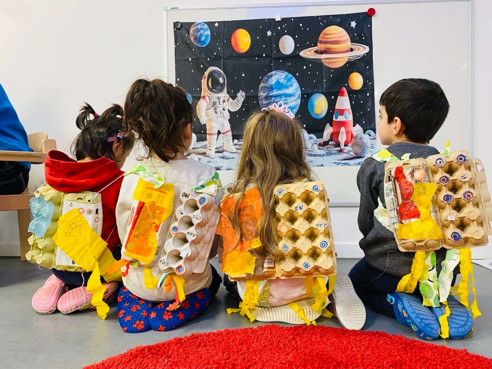 Kinder von hinten vor einem Planetenposter. Auf dem Rücken haben sie selbstgebastelte Antriebsraketen aus Eierkartons.