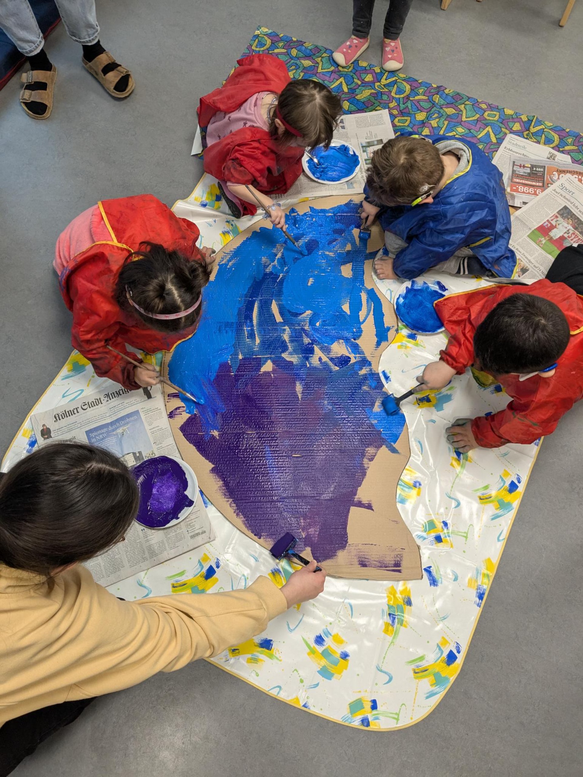 Von oben: Kinder malen auf dem Fußboden eine große Pappe mit blauer und lilafarbener Farbe an.