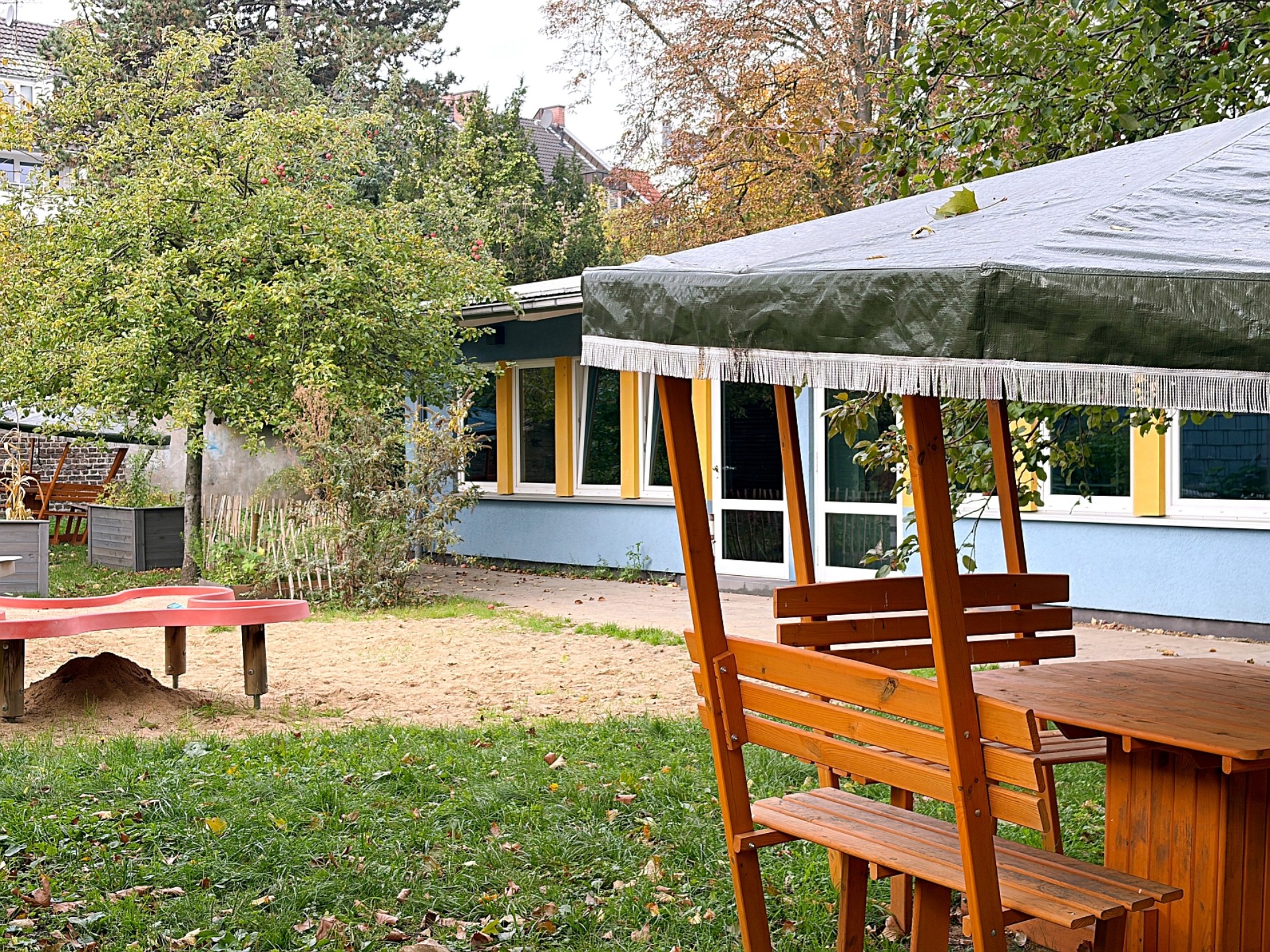Eine überdachte Tischgruppe im Schulgarten, im Hintergrund das blaue Haus der OGS.